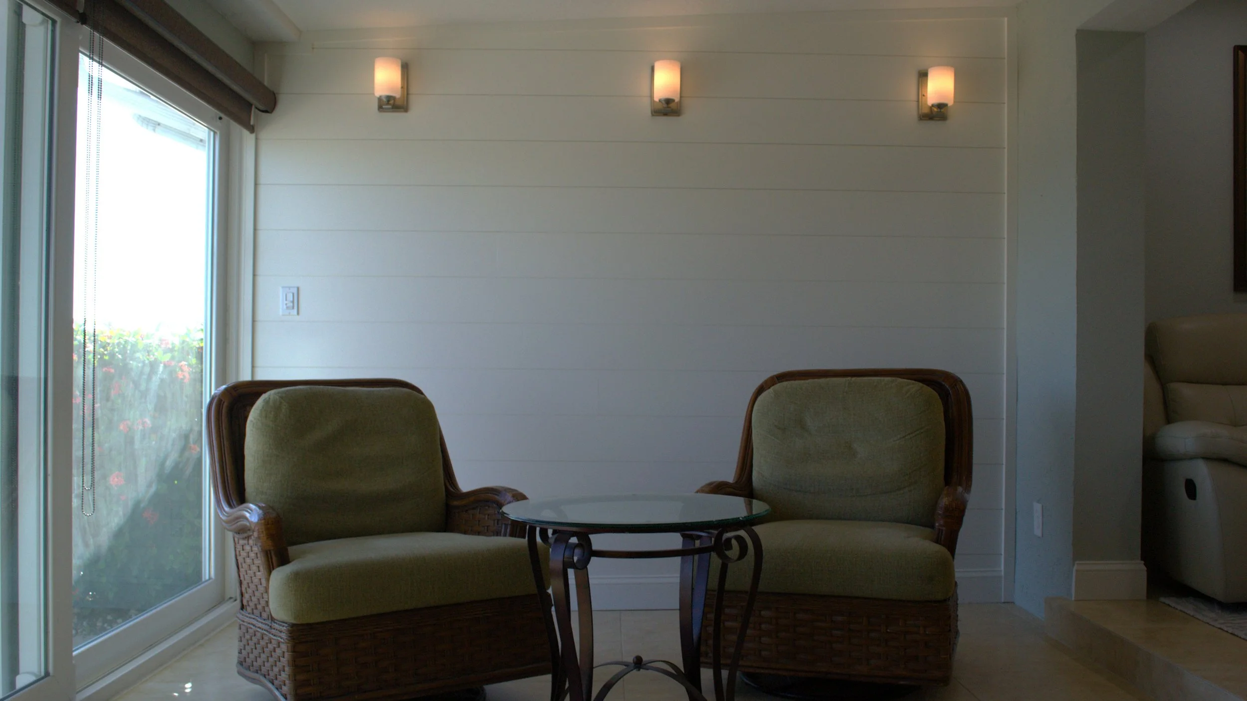 Living room with two wicker armchairs with green cushions, a small glass-top table between them, a large window on the left with a brown shade, and three wall-mounted lights above.