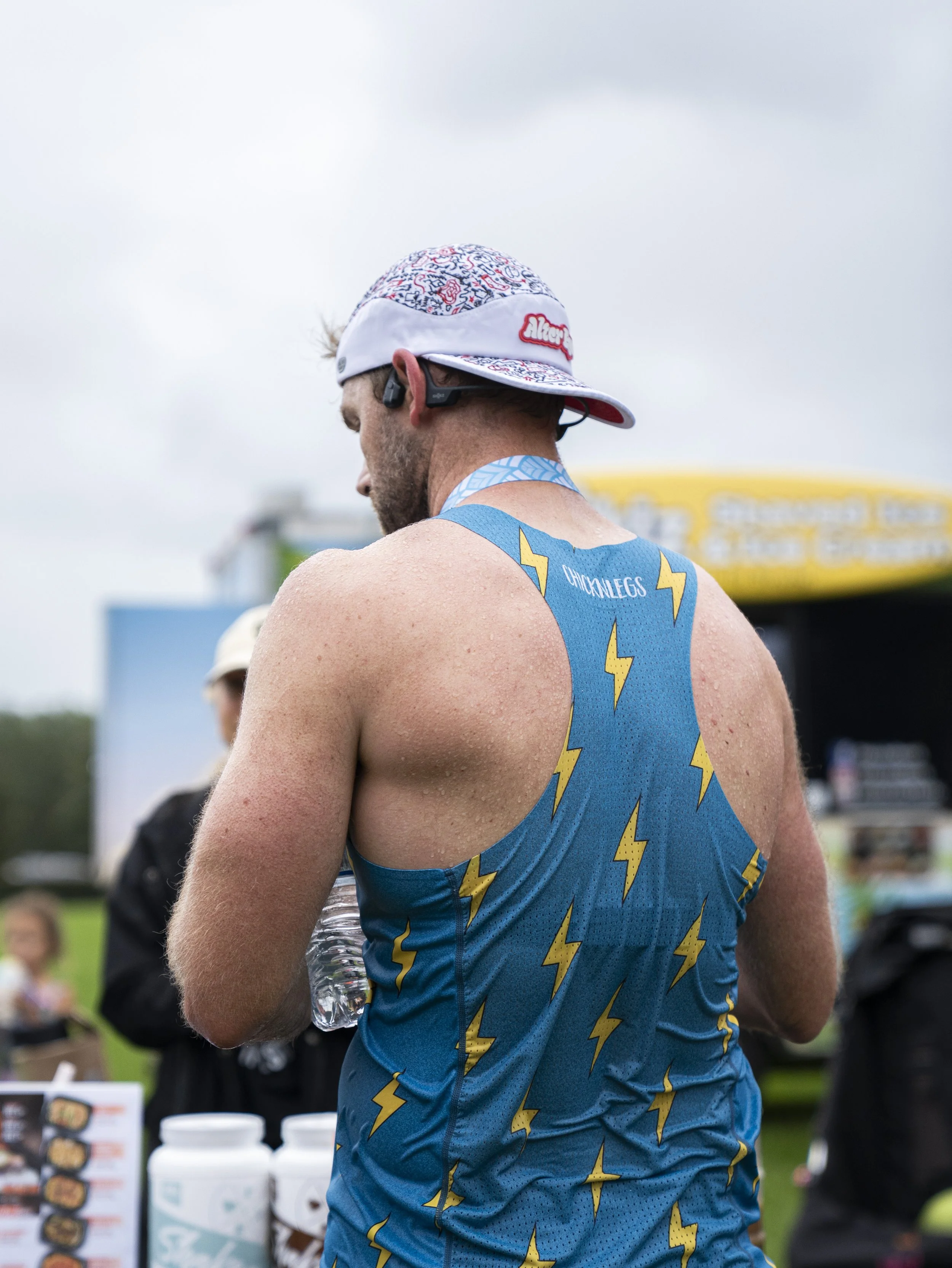 A man in a blue athletic tank top with yellow lightning bolts, wearing a cap and Bluetooth earpiece, standing outdoors during a race or event.