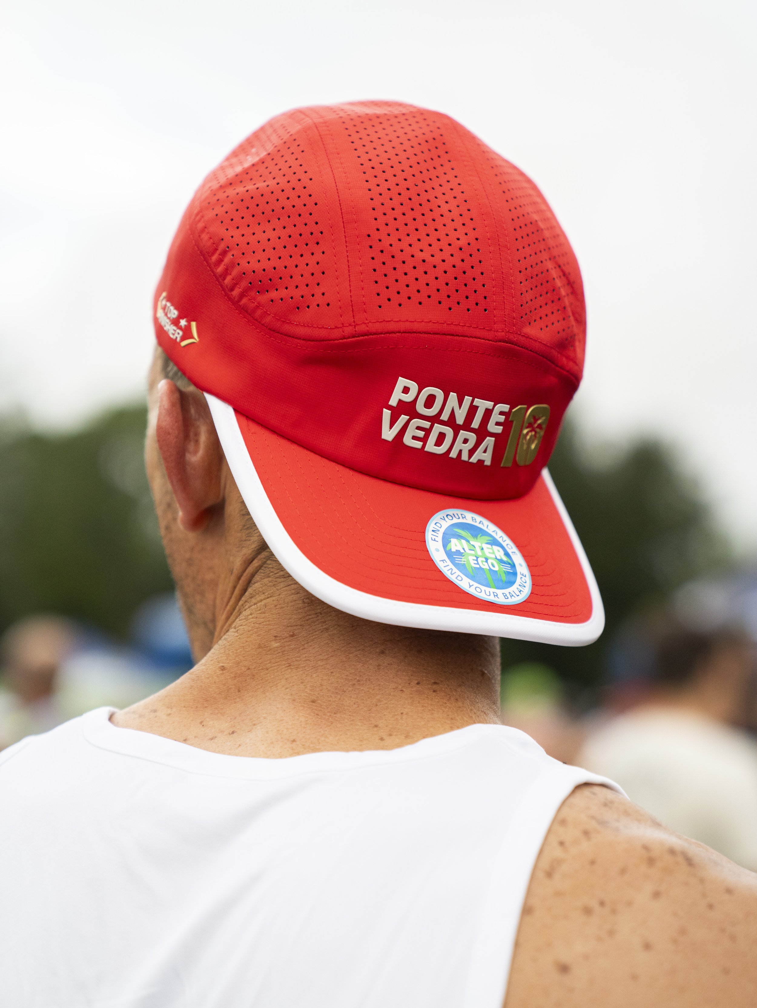 Back view of a man wearing a red cap with the words 'Ponte Vedra' and the number 10, and a white tank top, outdoors on a cloudy day.