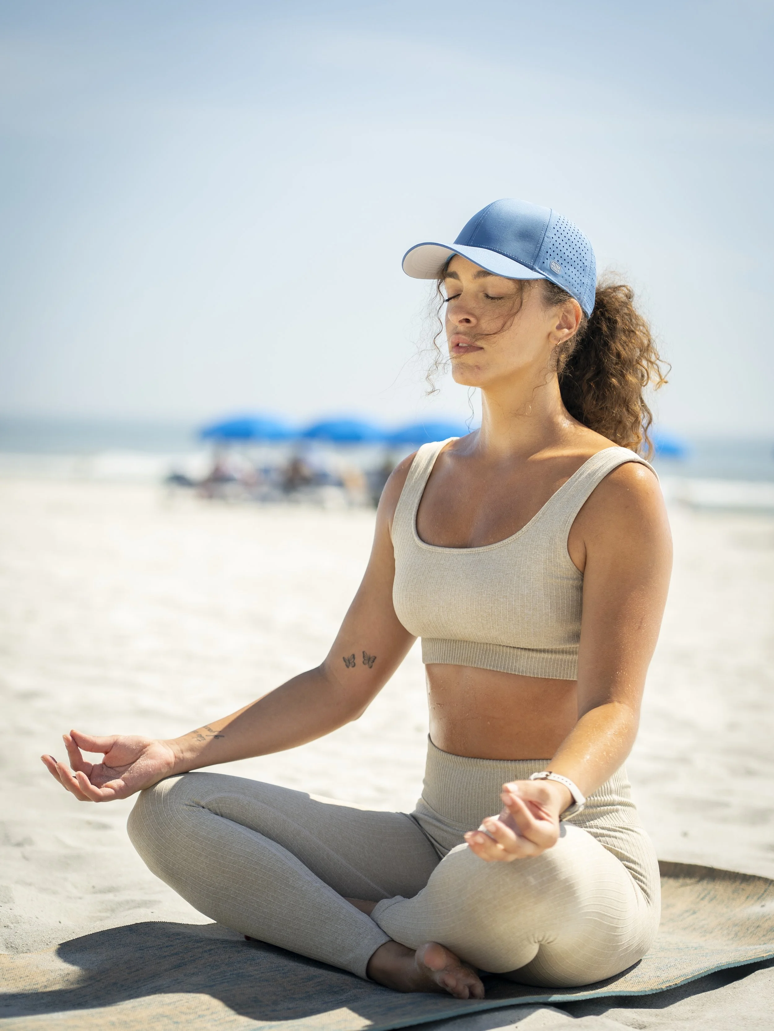 Woman practicing yoga on the beach, sitting cross-legged with eyes closed, wearing a beige sports bra, matching leggings, and a blue cap.