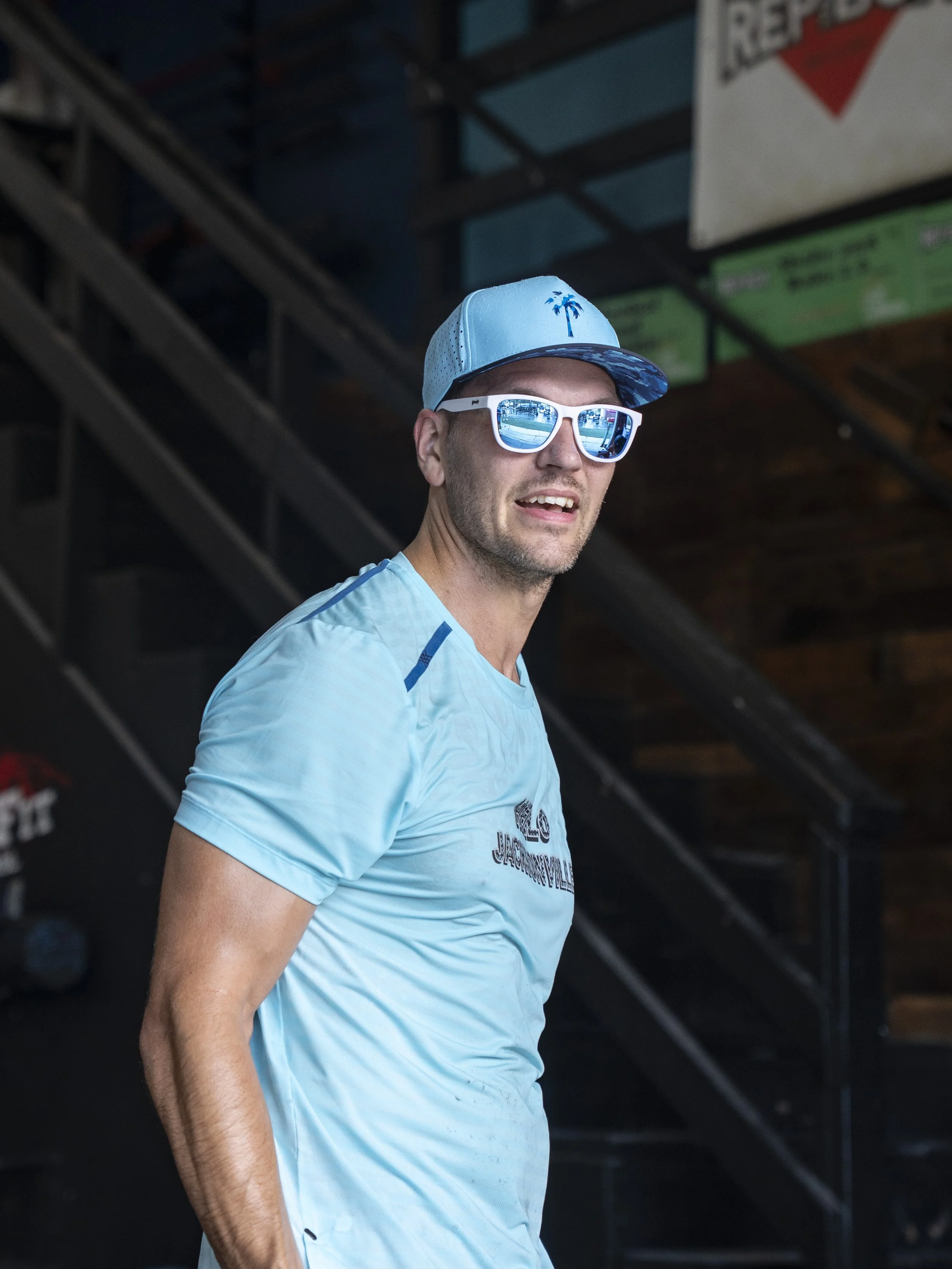 Man wearing light blue athletic shirt, white sunglasses, and a blue cap with a palm tree logo, standing indoors near stairs.