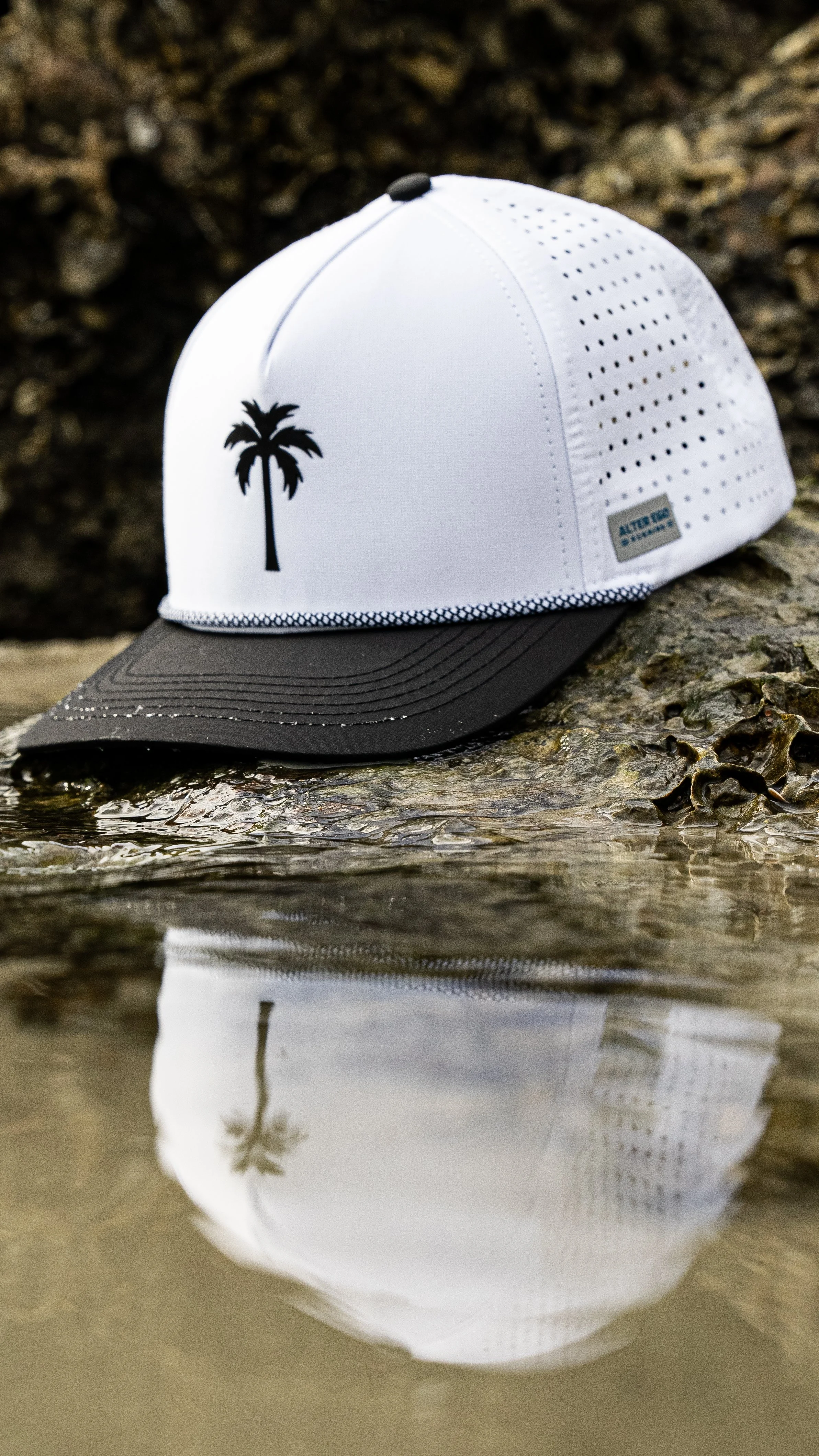 A white and black cap with a palm tree logo, resting on rocks and water, reflecting in the water.