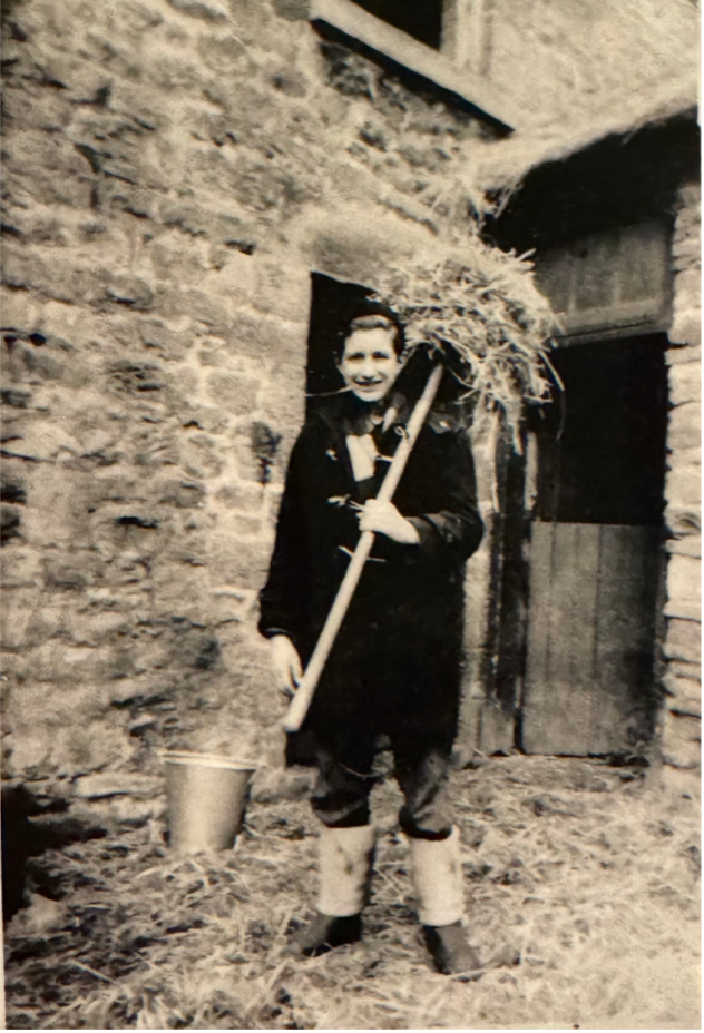 a sepia photo of John Meadley aged 17 standing in front of a farm building holding a pitch fork over his shoulder with hay on it.