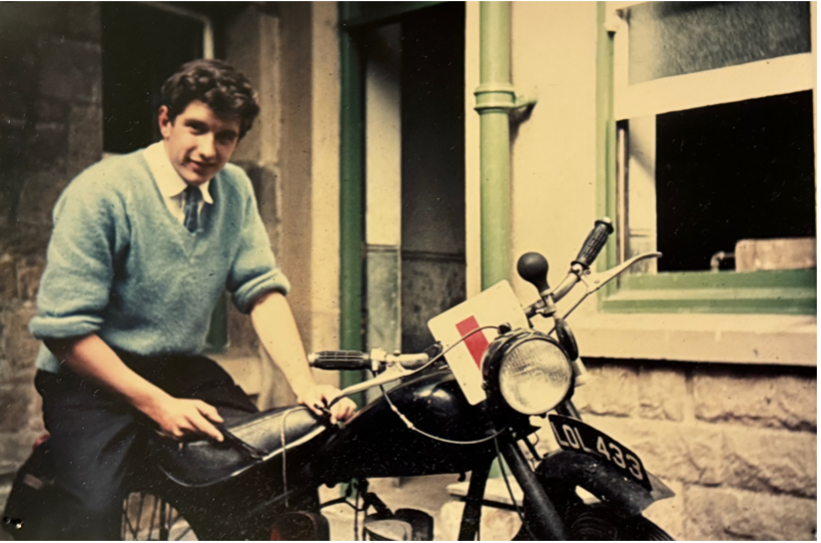 John Meadley as a young man sitting on a vintage motorcycle outside a stone building, with a green window and pipes behind him.