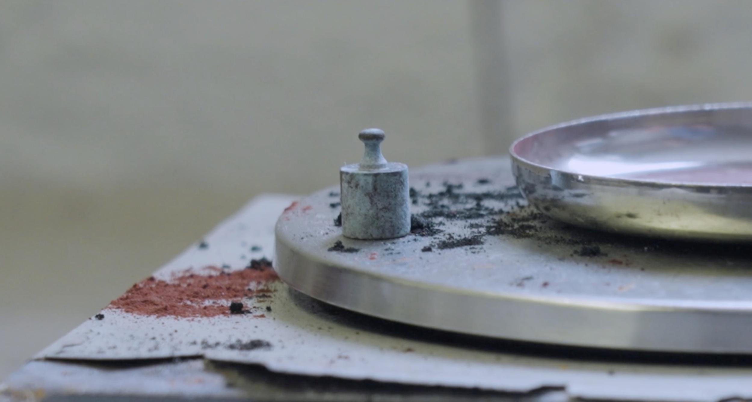 Close-up of a metal fixing on a surface next to a shallow metal dish with liquid, surrounded by colourful pigment