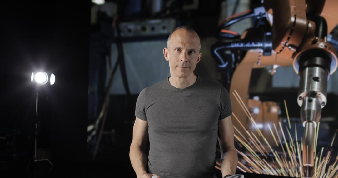A man in a gray T-shirt stands in front of robotic arms and sparks, in a high-tech industrial setting.