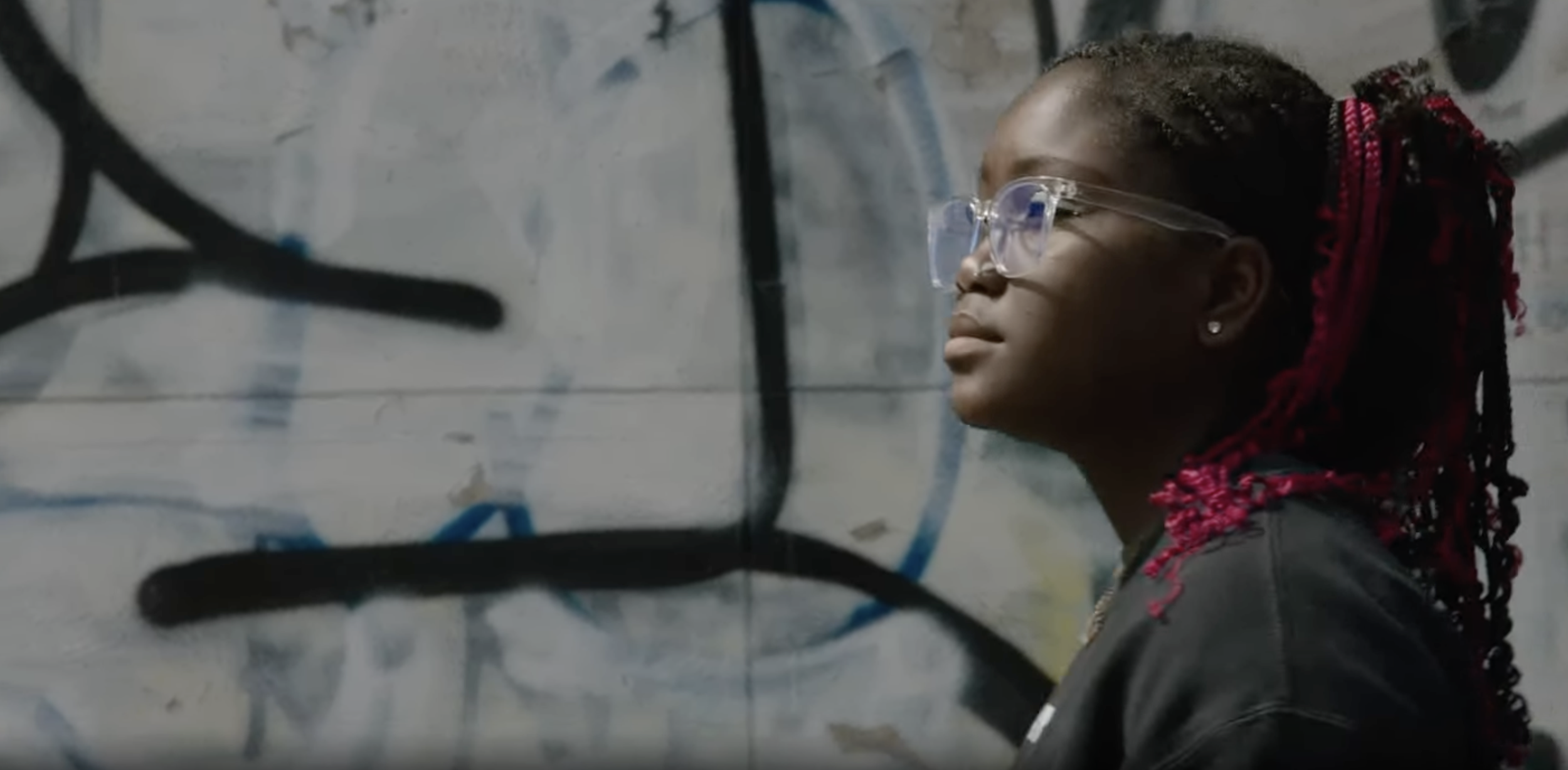 Side profile of a young woman with braided red hair, wearing glasses and earrings, standing against a graffiti-covered wall.