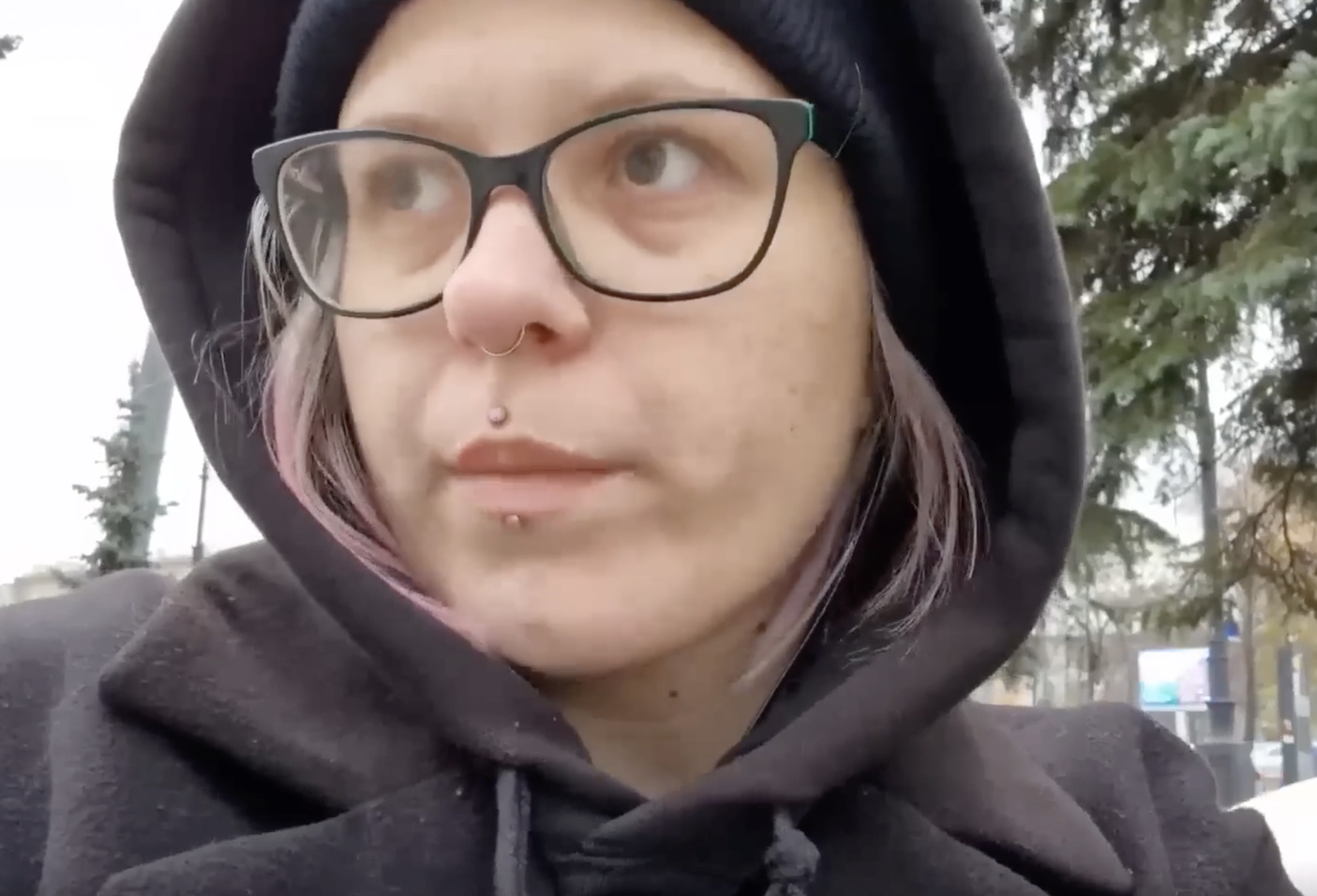 A person wearing glasses and a black hoodie outdoors, with trees and a cloudy sky in the background.