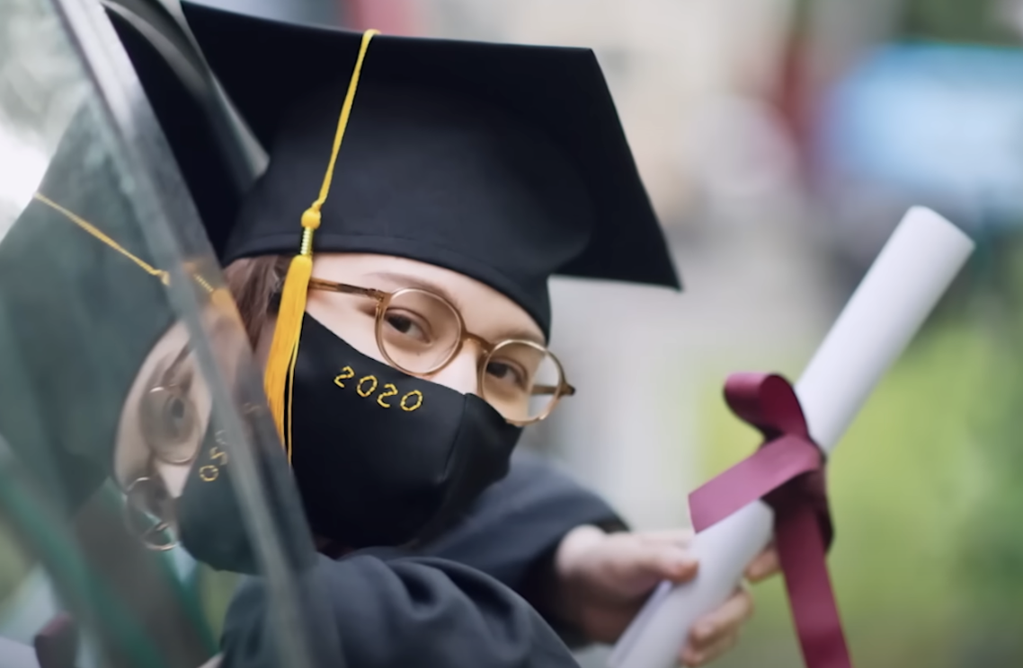 A young graduate wearing a black cap, gown, and face mask with '2020' written on it, holding a rolled diploma tied with a purple ribbon.