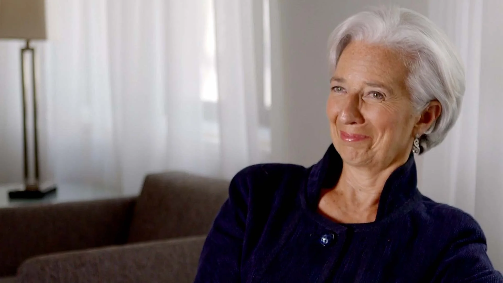 Christine La Garde wearing a dark top, sitting on a sofa in a room with white curtains and a lamp in the background.
