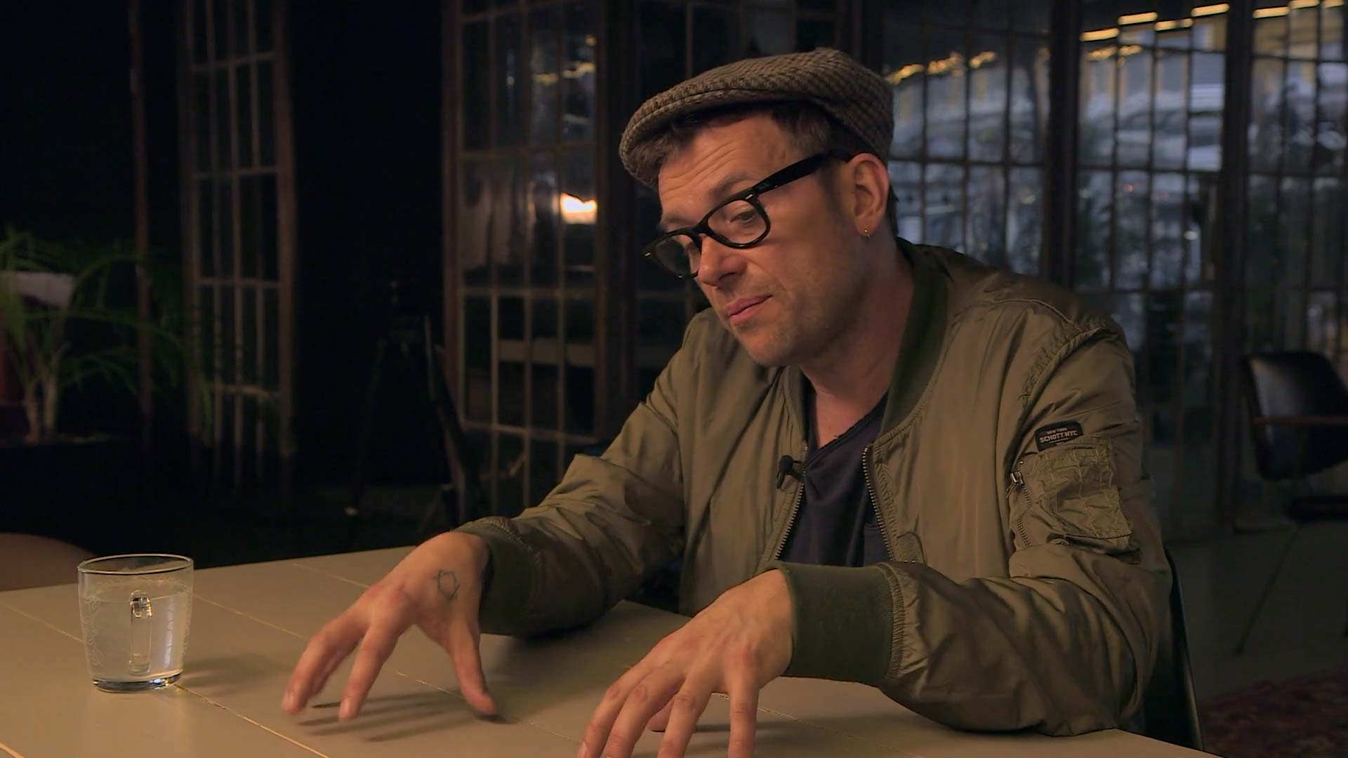 Damon Albarn wearing glasses, a green jacket, and a flat cap is sitting at a table with a glass of water, in a dimly lit room with large windows behind him.