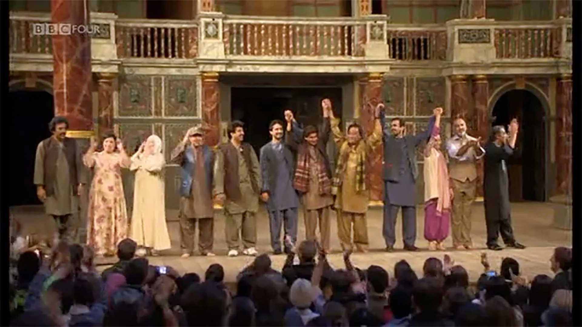 The cast of a theatrical performance stands on stage, holding hands and taking a bow in front of an audience, in a theater with ornate, colorful decor.