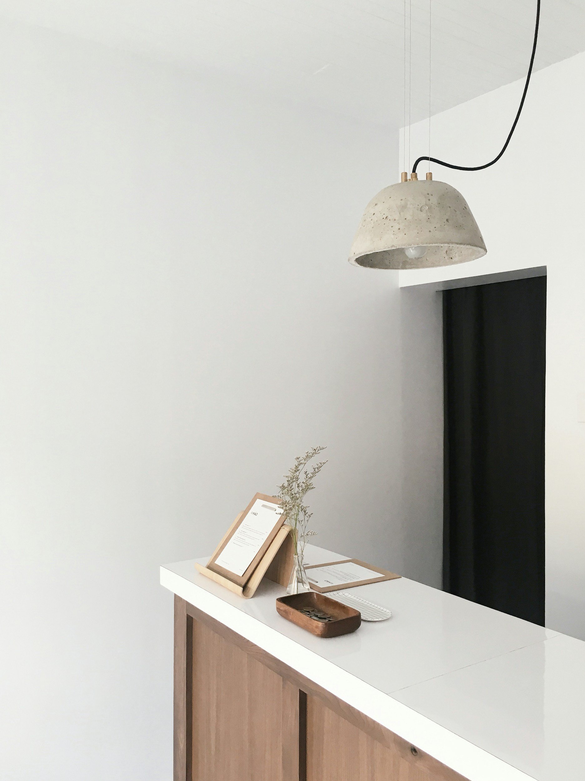 Minimalist reception desk with a small clear vase holding dried green branches, a framed menu or sign, a wooden tray, and a white keyboard, with a concrete ceiling light hanging above. The decor is modern and simple, with neutral tones.