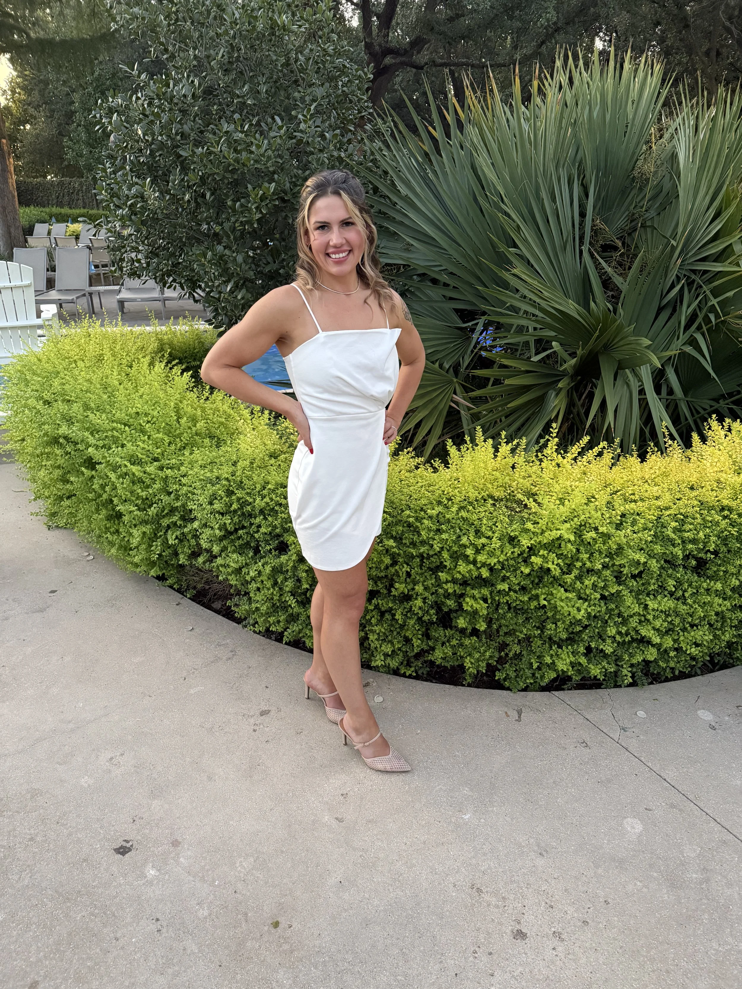 A woman in a white dress and pink high heels standing outdoors in front of green bushes and large plants, smiling at the camera.