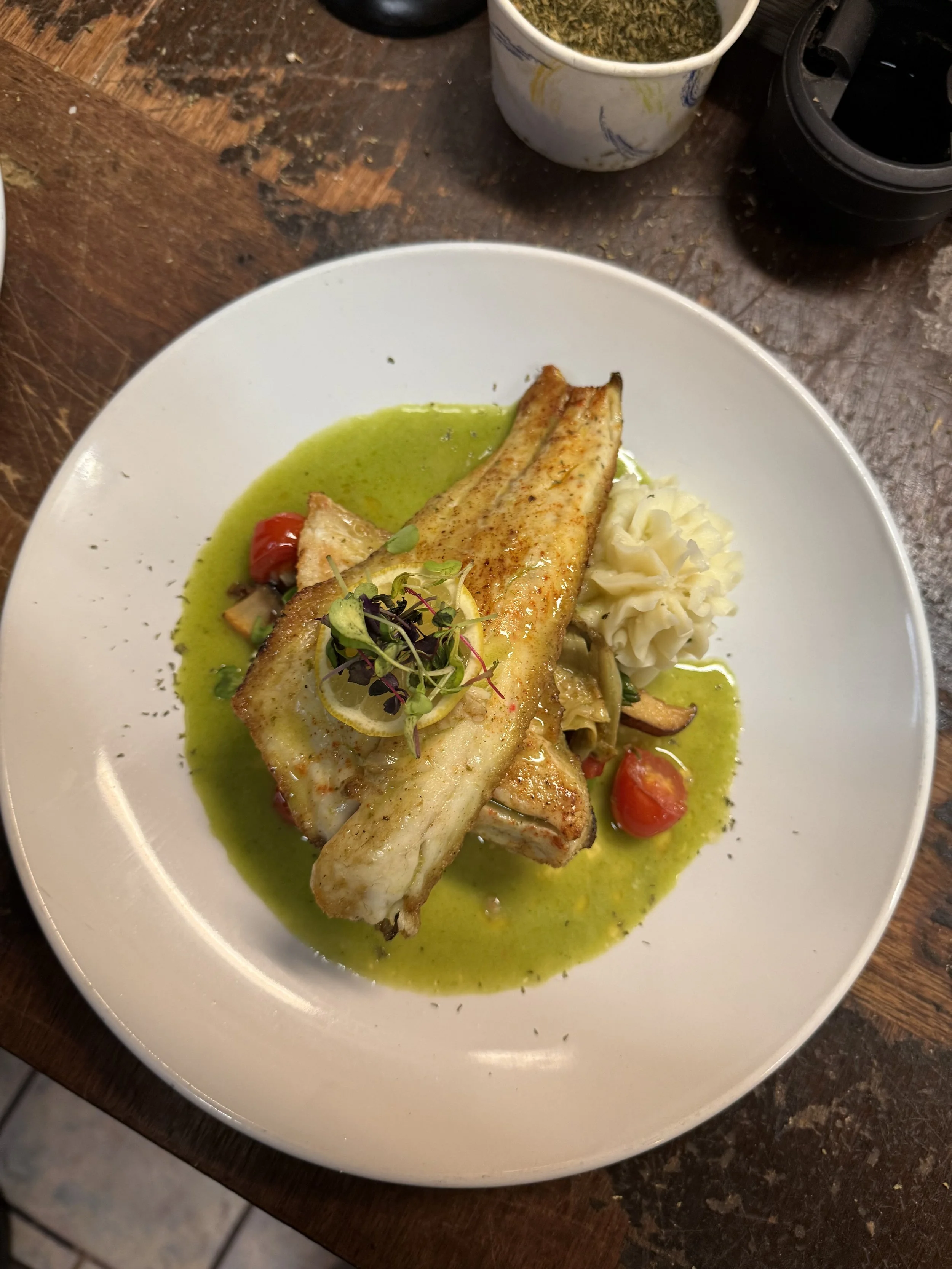 A plated dish featuring grilled fish with a green sauce, accompanied by mashed potatoes, cherry tomatoes, and garnished with microgreens.