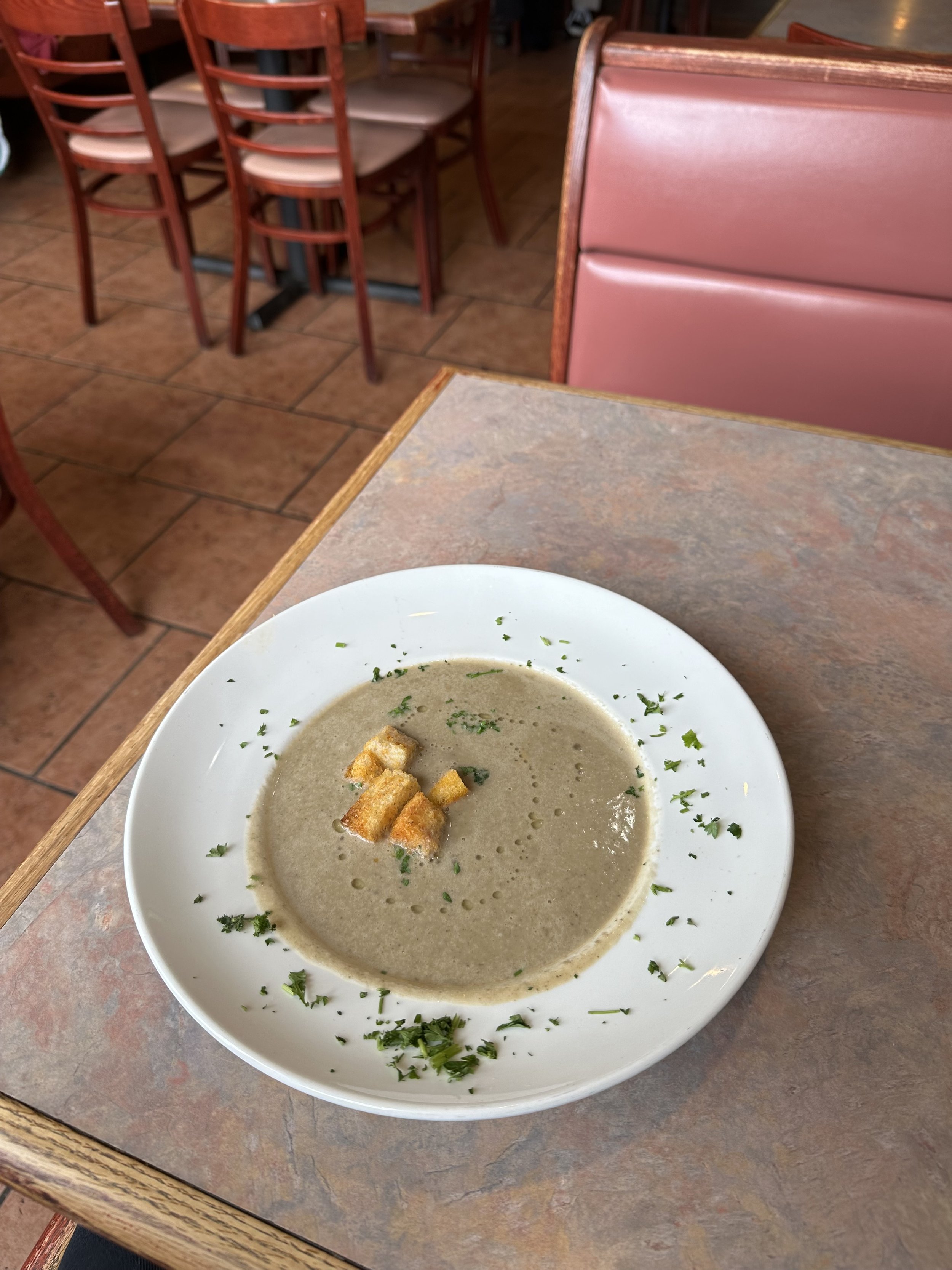 Bowl of creamy mushroom soup garnished with chopped parsley and croutons, on a wooden table at a restaurant.