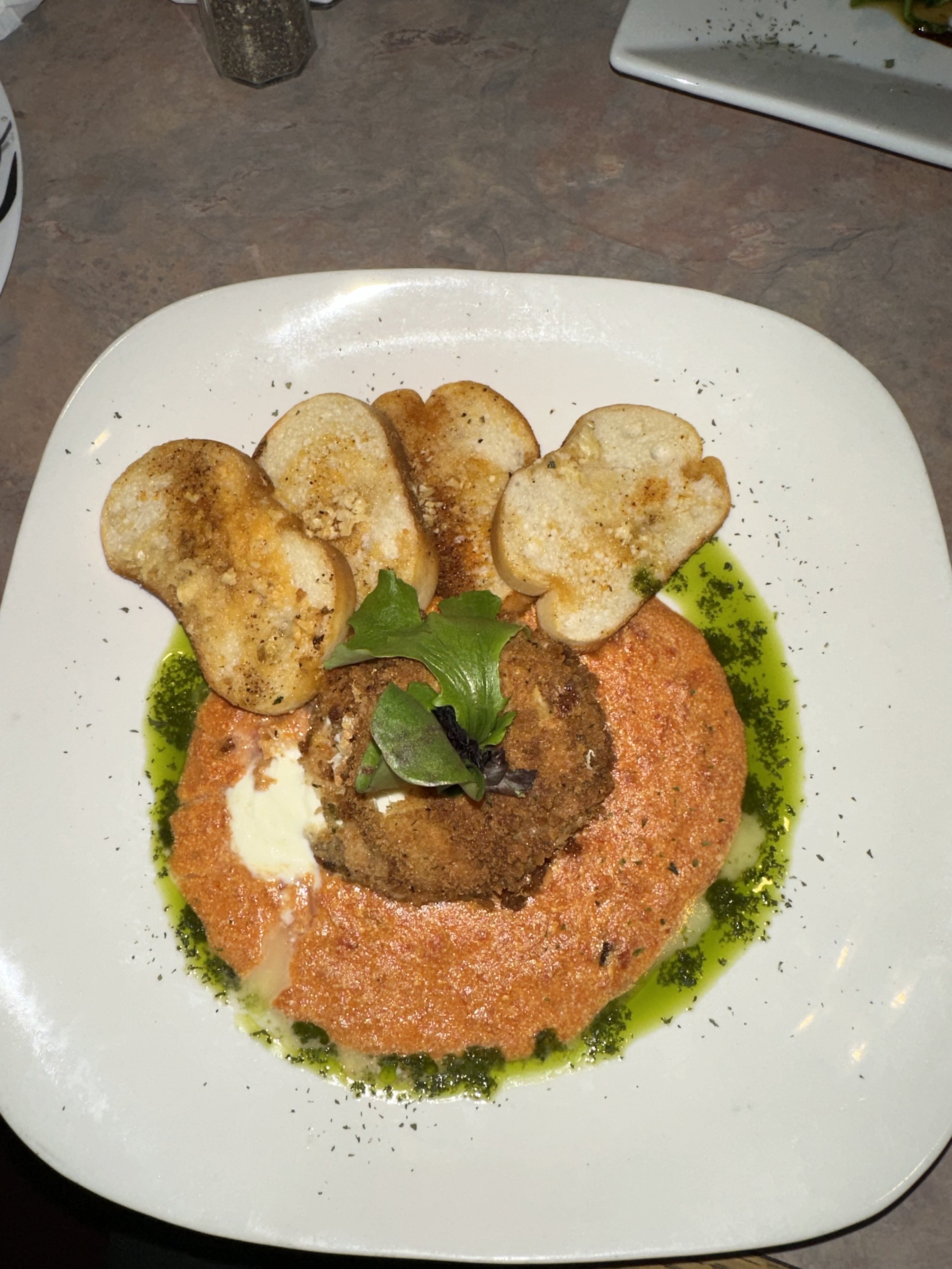 A plated serving of veal Milanese with mashed potatoes, topped with a fried meat patty and garnished with greens, surrounded by green oil or sauce, on a white square plate.