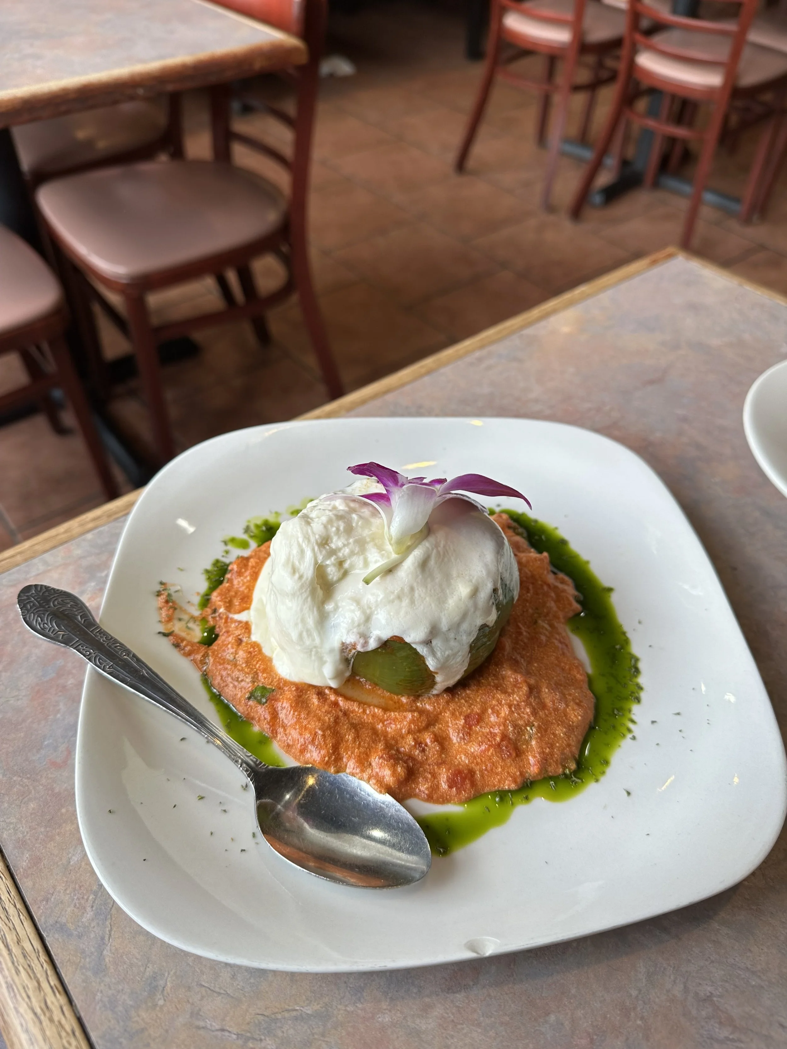 Plate of food with a scoop of white creamy substance topped with an edible flower, on a bed of green sauce, served on a white square plate.
