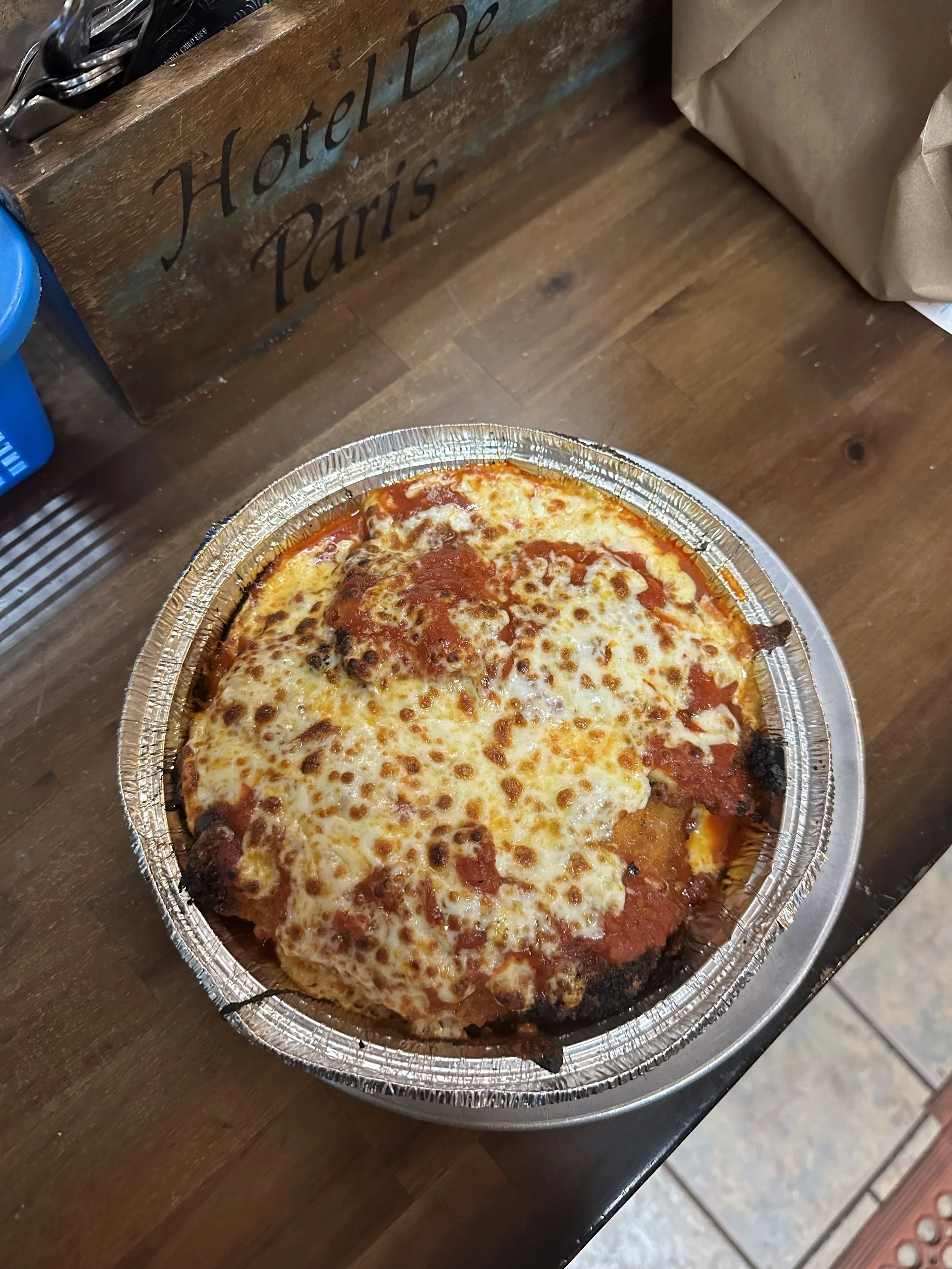 Personal-sized cheese pizza in an aluminum takeout container on a wooden table.