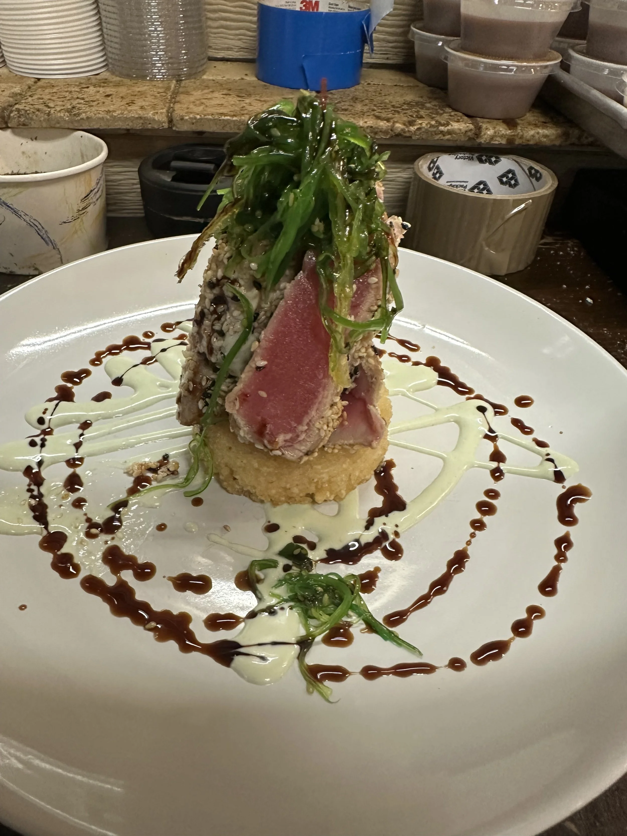 A plated gourmet dish featuring seared tuna with crispy coating, topped with green vegetables and garnished with drizzles of white and dark sauces, served on a large white plate.