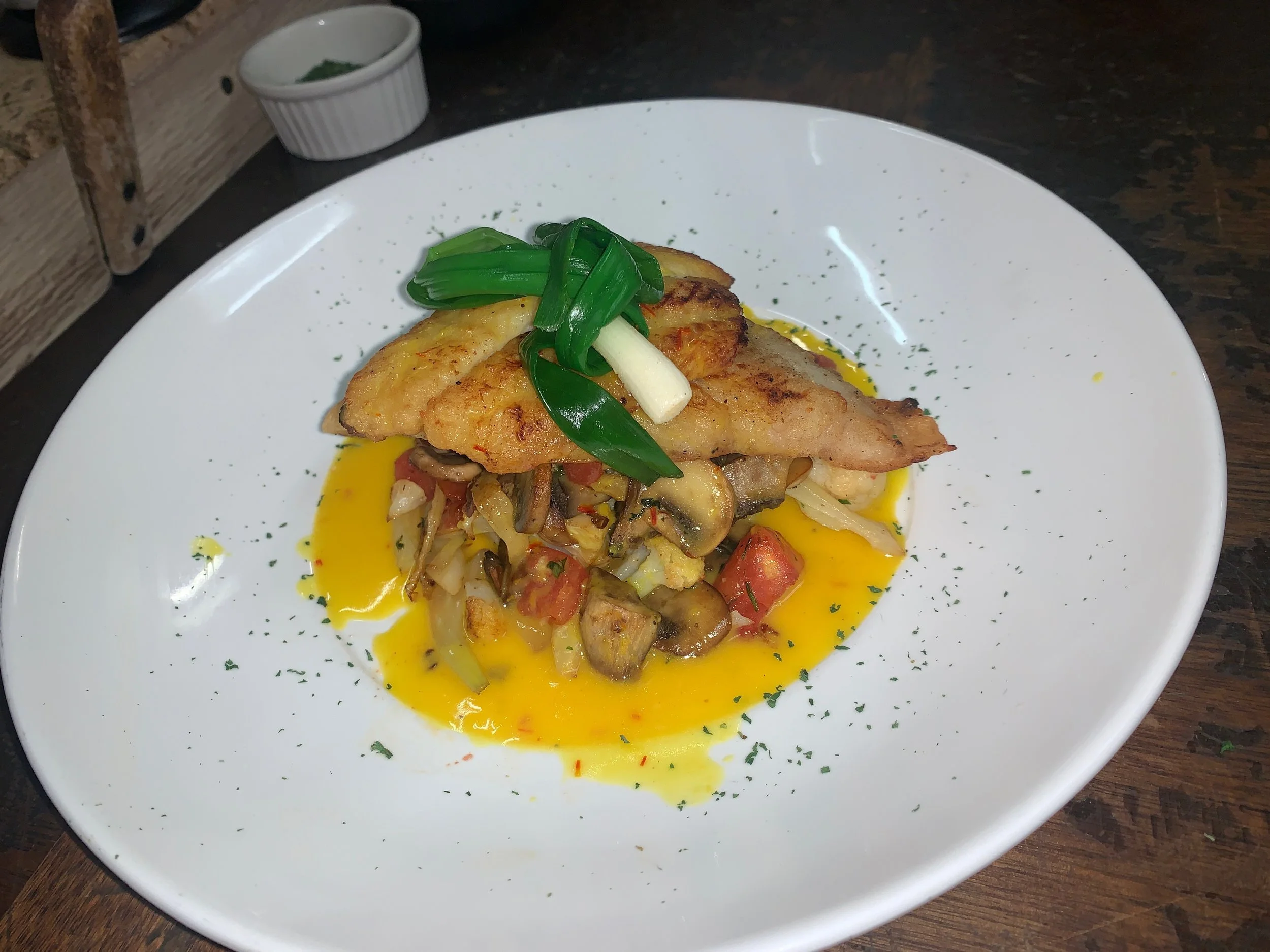 Plate of grilled fish with sautéed vegetables, garnished with green onions and herbs, served with a yellow sauce.