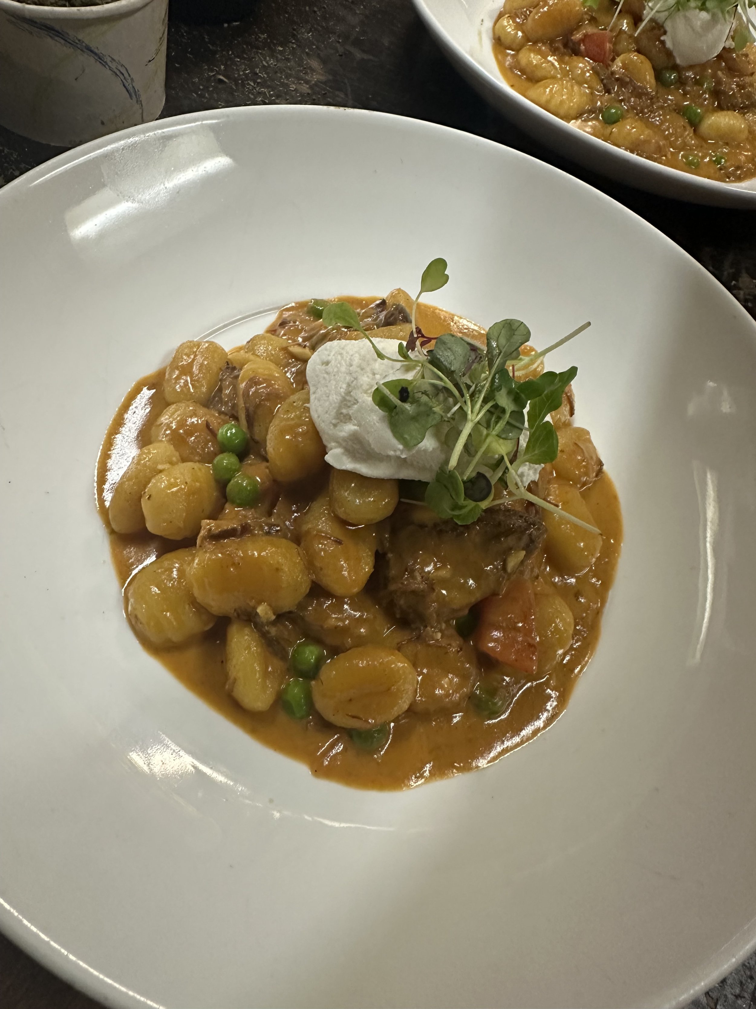 A plate of gnocchi with a creamy orange sauce, garnished with microgreens and a dollop of white cream, with a bowl of more gnocchi in the background.