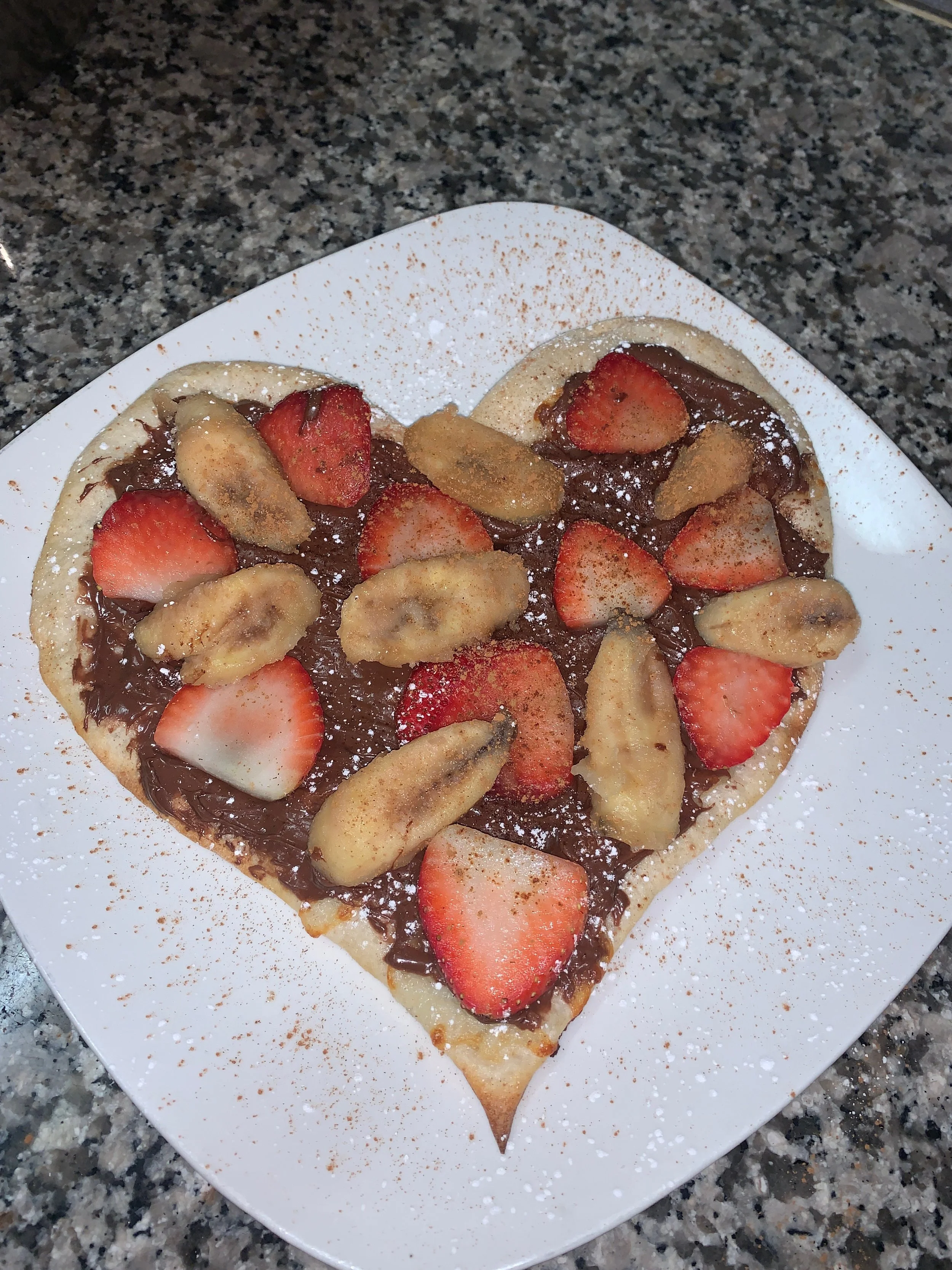 Heart-shaped waffle topped with sliced strawberries, banana slices, chocolate spread, and dusted with powdered sugar, served on a white plate on a granite countertop.