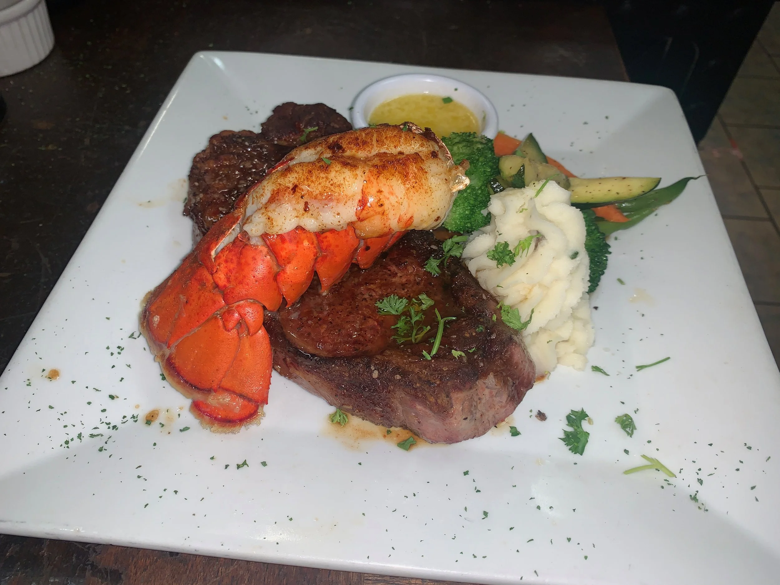 A plate of steak with mashed potatoes, lobster, vegetables, and a small bowl of sauce, garnished with herbs.