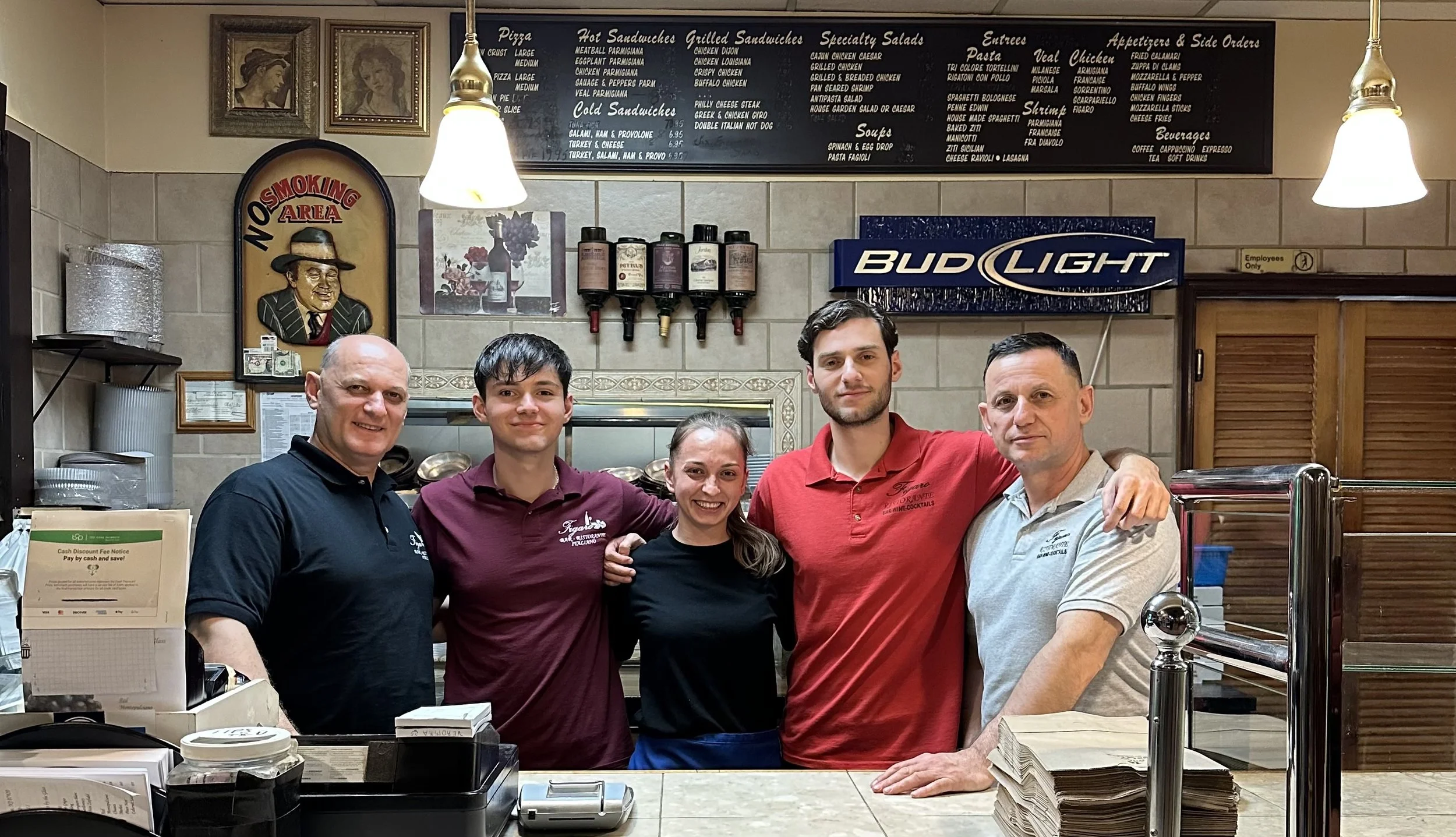 Five restaurant staff members stand behind the counter, smiling, with a menu board and restaurant decor in the background.