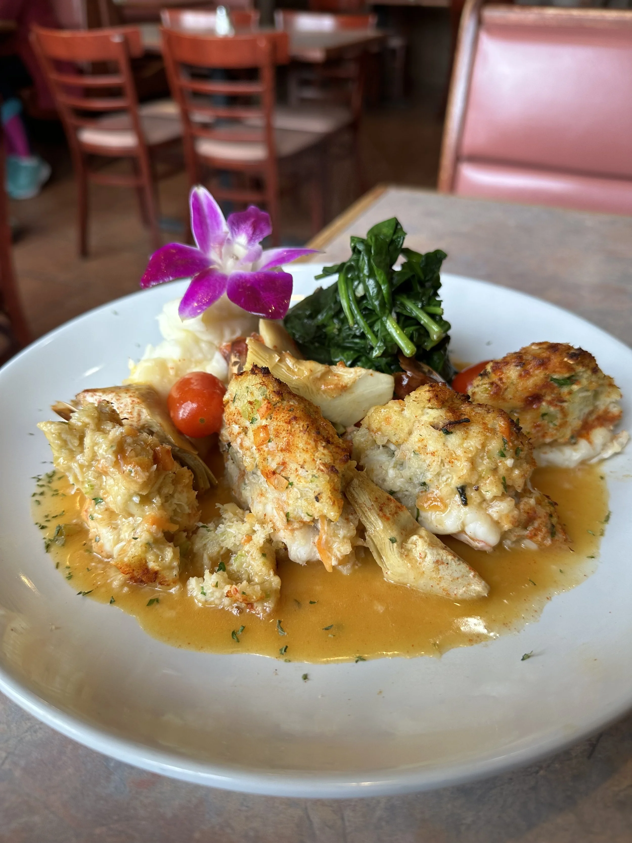 Plate of crab cakes garnished with herb sauce, served with mashed potatoes, sautéed spinach, a cherry tomato, and a purple orchid flower on top, in a restaurant setting.