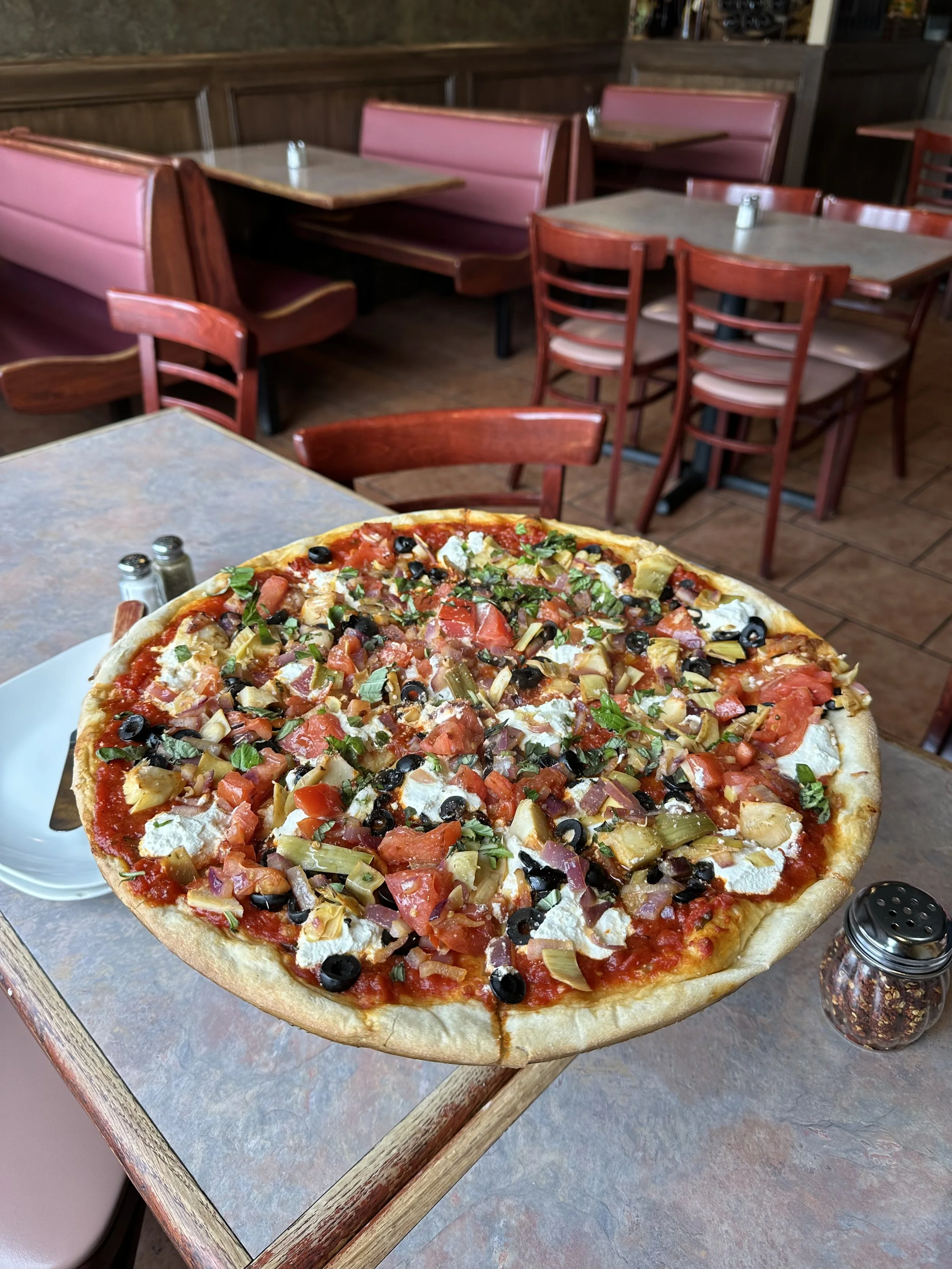 A pizza topped with tomatoes, black olives, green peppers, onions, basil, and cheese on a wooden tray in a restaurant with tables and chairs in the background.