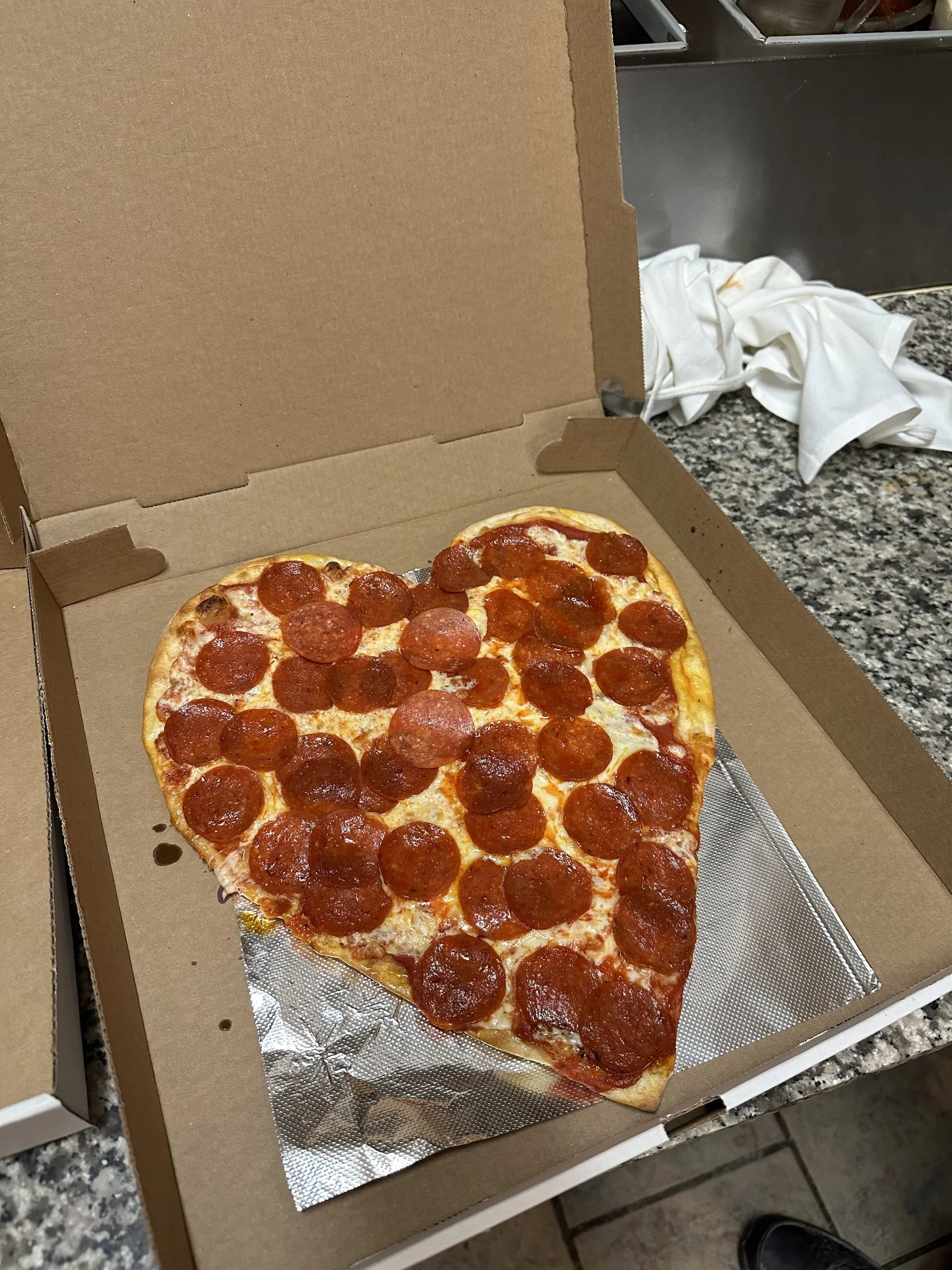 Heart-shaped pepperoni pizza in an open cardboard box on a granite countertop.
