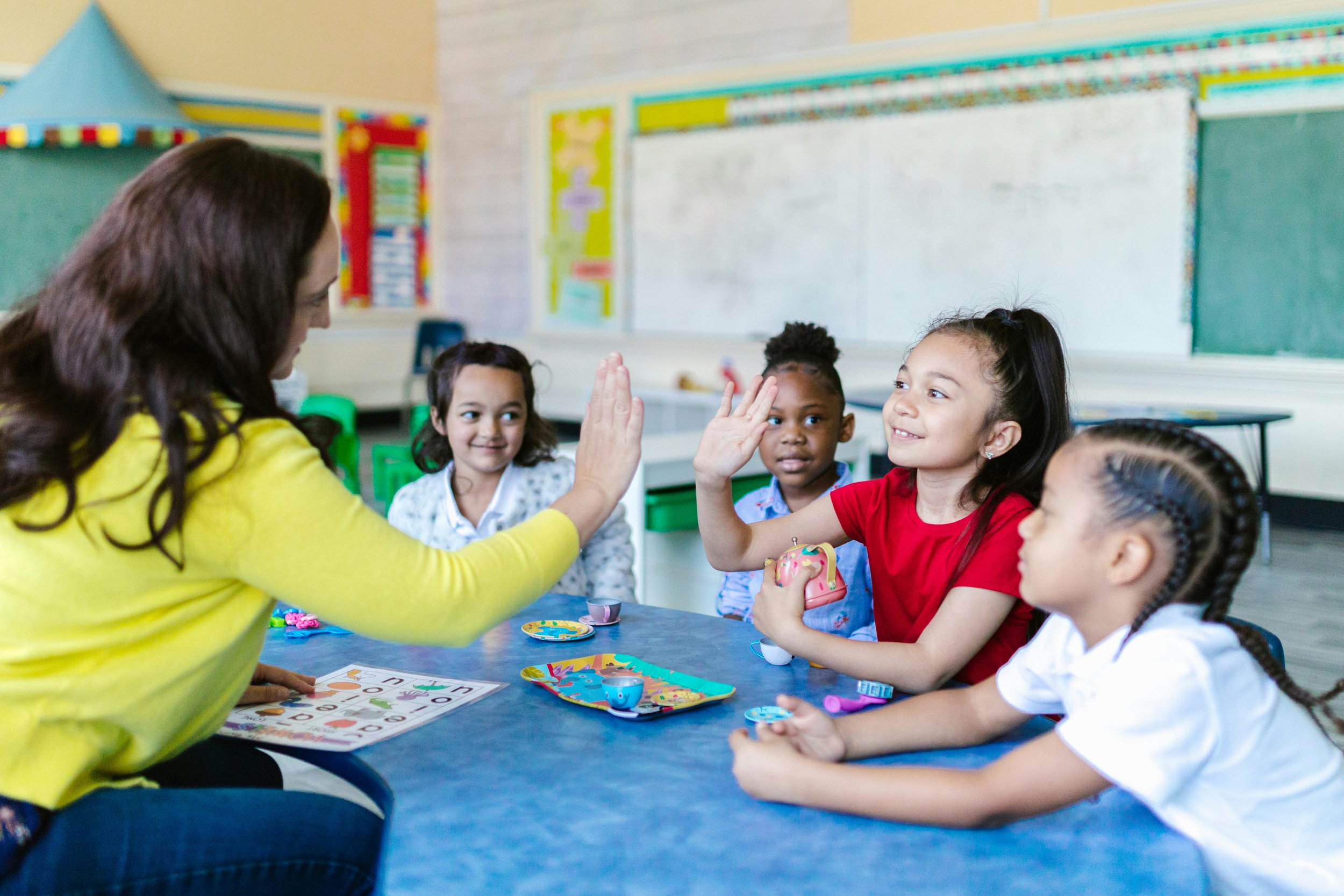 A teacher giving a high five to a girl in a red shirt during a classroom game with four young students sitting at a table with toys and game pieces.