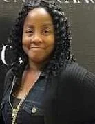 Woman with curly hair, smiling, wearing a black jacket and a pearl necklace