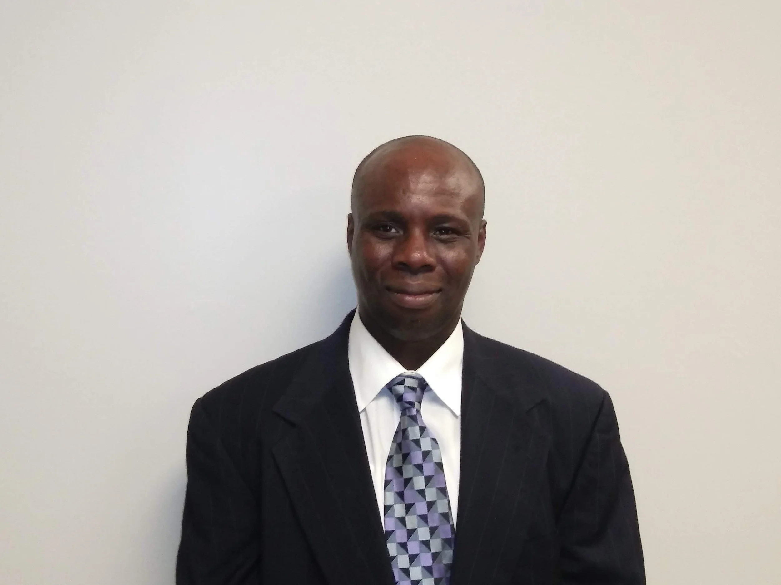 A man in a dark suit, white shirt, and patterned tie standing against a plain white wall.