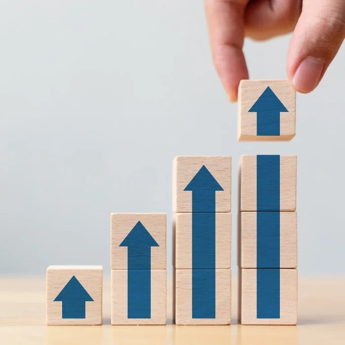 Hand stacking wooden blocks with upward blue arrows, forming a rising bar graph to show growth or progress.