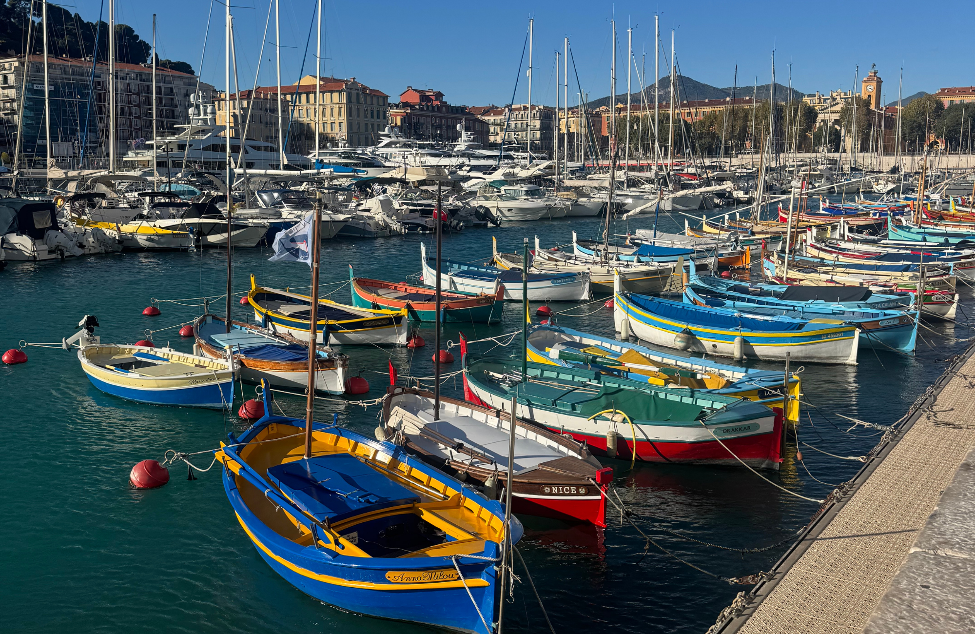 Colorful boats docked at a marina with buildings and mountains in the background.