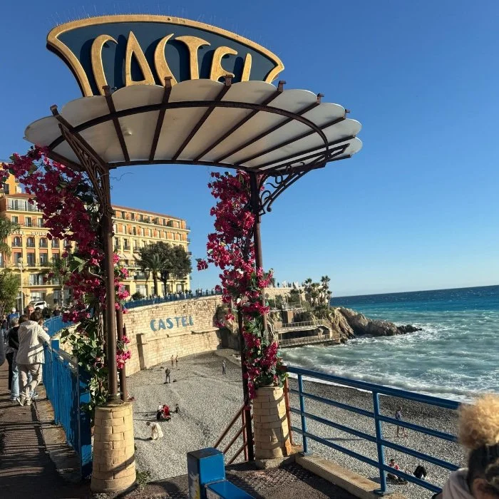 A seaside walkway with a Castel sign, pink flowers, and people on a rocky beach by the blue Mediterranean Sea under a clear sky.