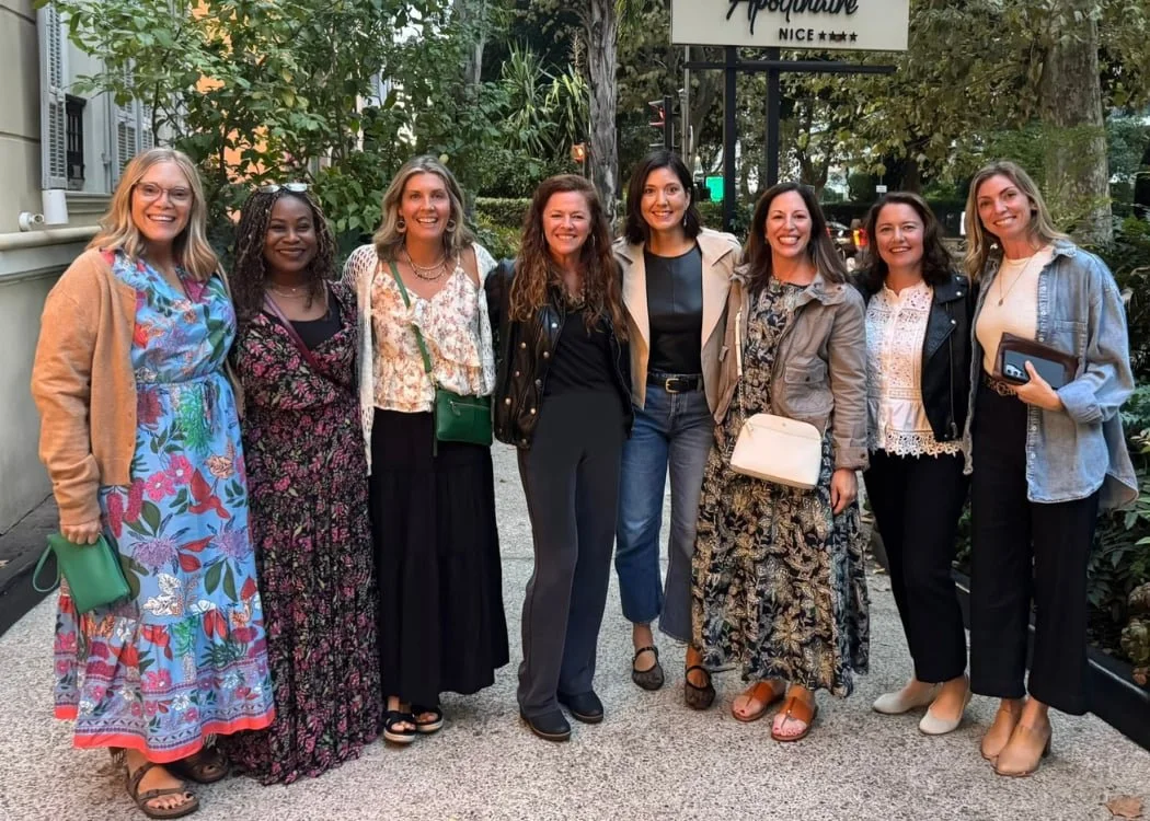 Eight women stand together outdoors, smiling for a group photo, dressed in colorful, casual clothing with greenery in the background.