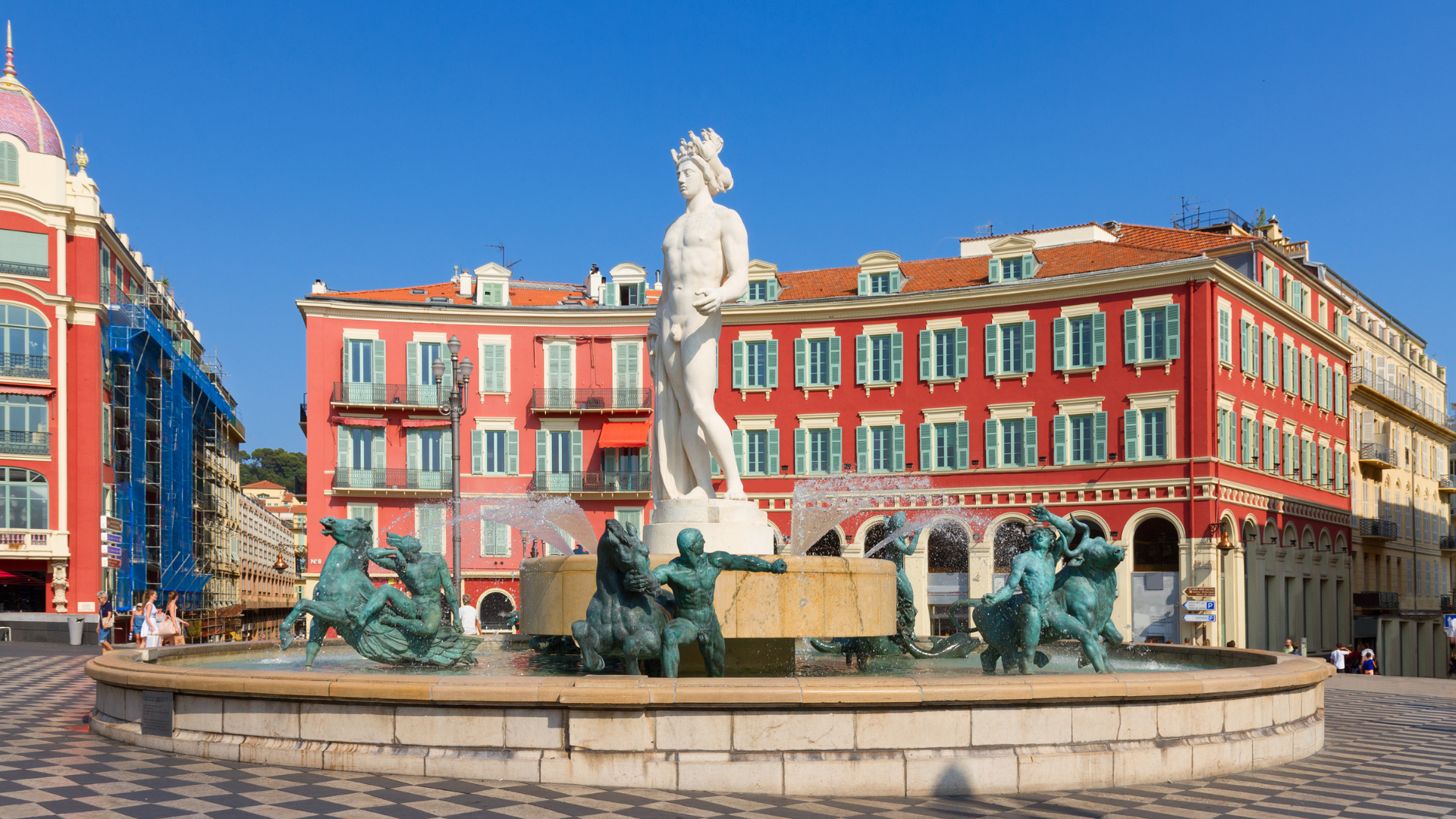 Colorful city square with a fountain featuring a classical statue of a woman at the center, surrounded by bronze sculptures of mythological figures, with multi-story buildings with green shutters and red roofs in the background under a clear blue sky