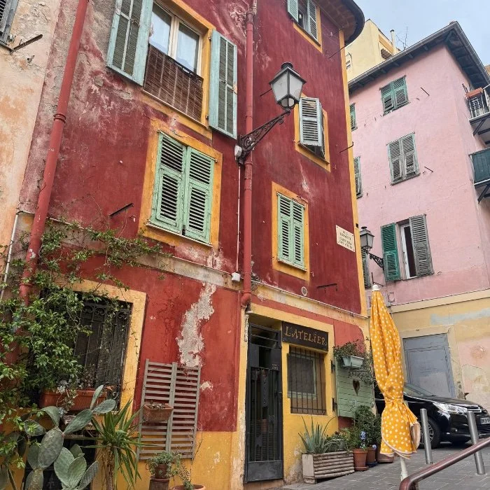 Colorful, weathered buildings with green shutters; yellow polka-dot umbrella and potted plants line a quaint, narrow street.