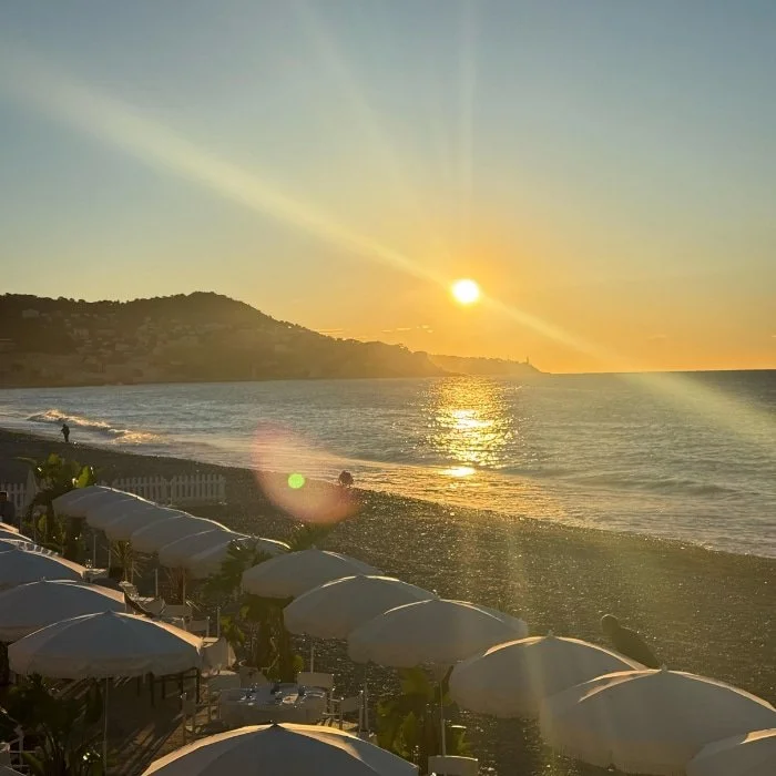 Sunset over a pebbled beach with white umbrellas, calm waves, and hills in the background.