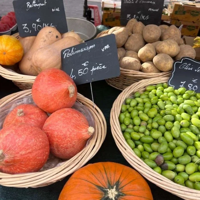 Baskets of squash, potatoes, and green olives at a market stall with handwritten price signs in French.