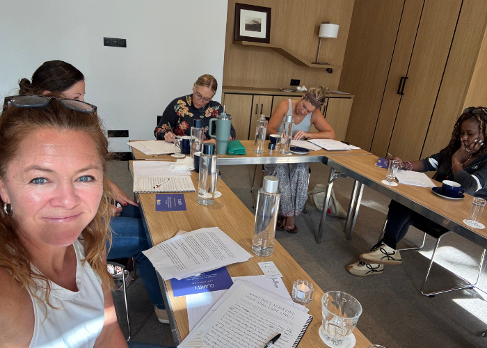 Five women sitting around a U-shaped conference table with notebooks, pens, water bottles, glasses, and documents; one woman is taking a selfie, smiling at the camera, in a well-lit meeting room with wood-paneled walls.