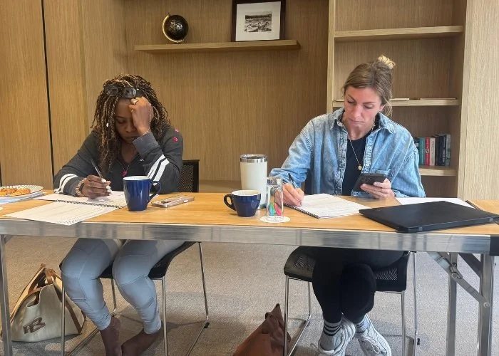 Two women sit at a table writing in notebooks; one looks at her phone, the other rests her head on her hand, both focused.