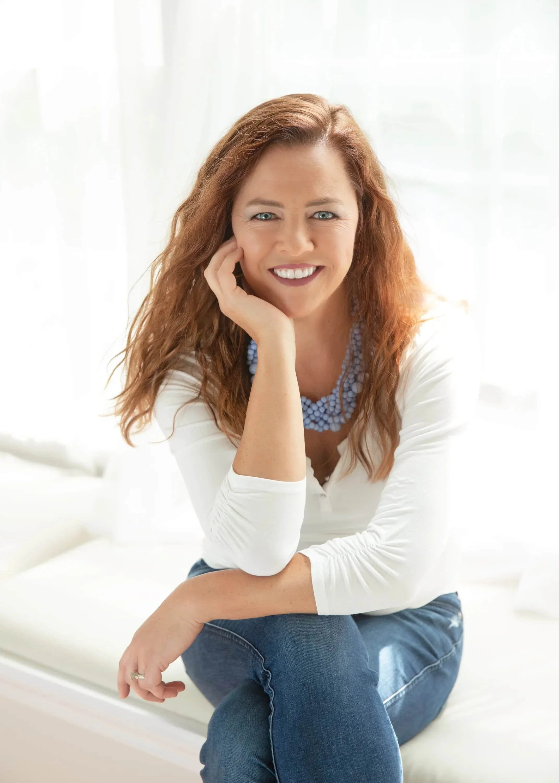 Woman with long red hair, wearing a white top and blue necklace, sits smiling with hand on her face in bright, natural light.