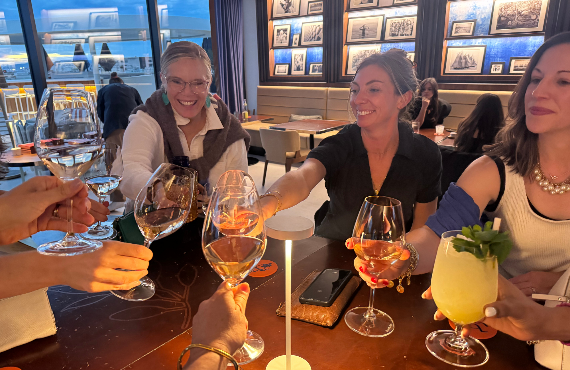 A group of women enjoying drinks at a restaurant, clinking glasses for a toast, with a lively atmosphere and wall art in the background.