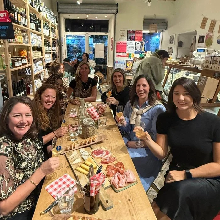 Seven women sit around a wooden table in a cozy deli, smiling and enjoying wine, charcuterie, and bread.