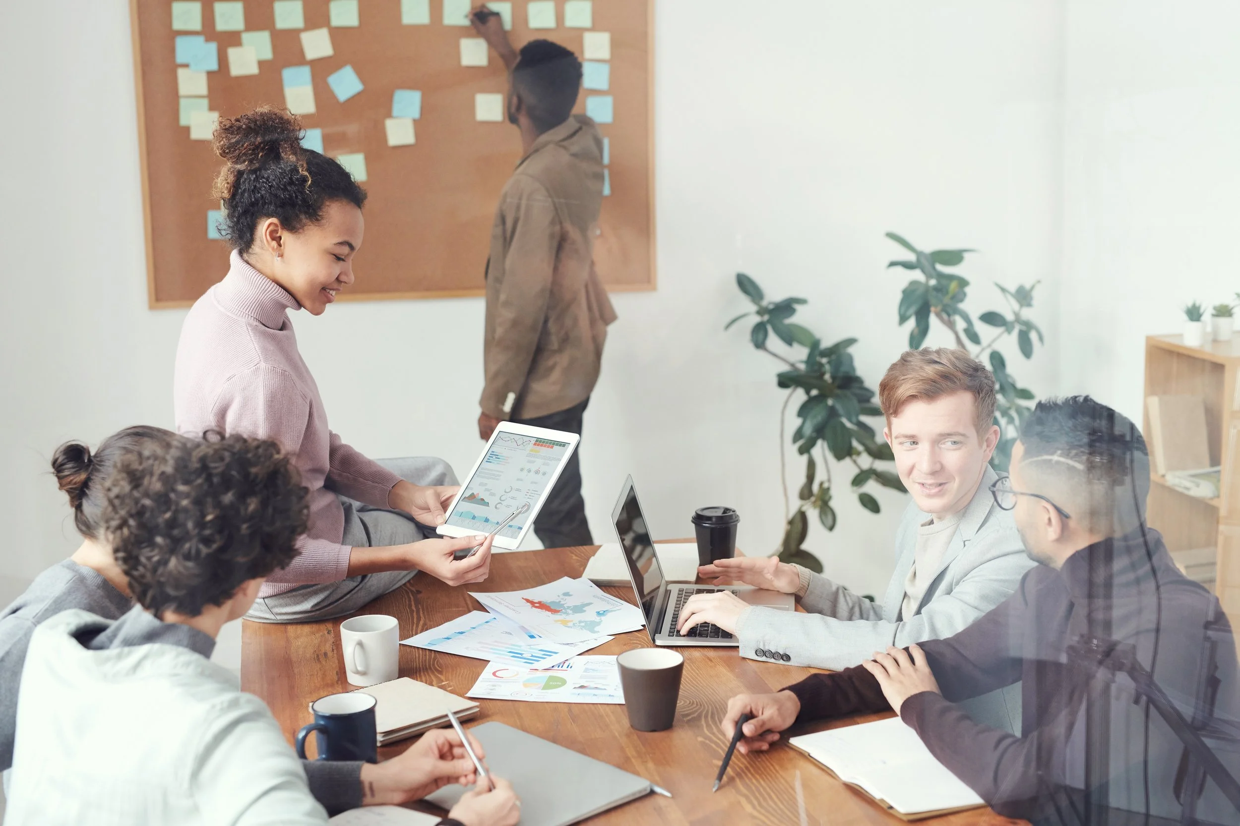 Four people in a meeting room, one woman sharing information from a tablet, three men listening and talking, documents and laptops on the table, a large plant in the background, and a man in the background writing on a corkboard with notes.