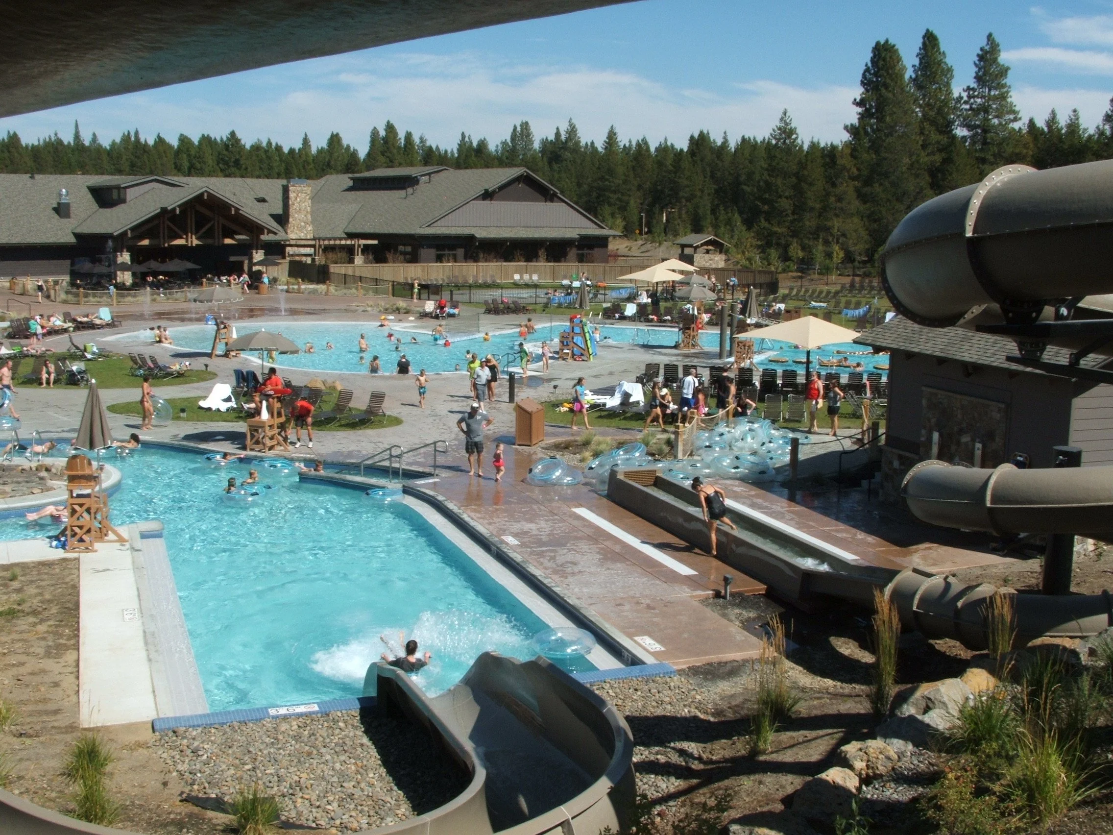 People enjoying a day at an outdoor water park with pools, slides, and lounge chairs, surrounded by trees and a mountain style building.